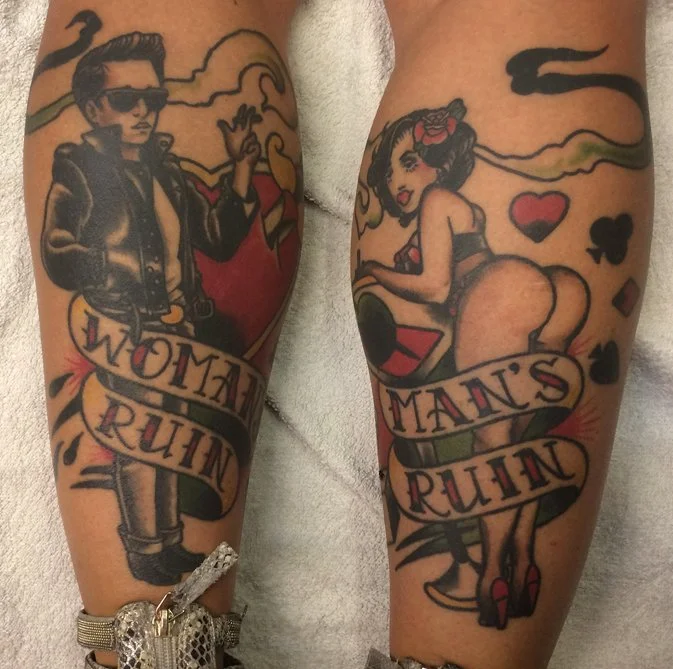 Tattooed legs featuring a man in sunglasses and leather jacket with a banner reading 'Woman's Ruin,' and a woman in revealing clothing with a banner reading 'Man's Ruin,' surrounded by playing card suits and a snake.