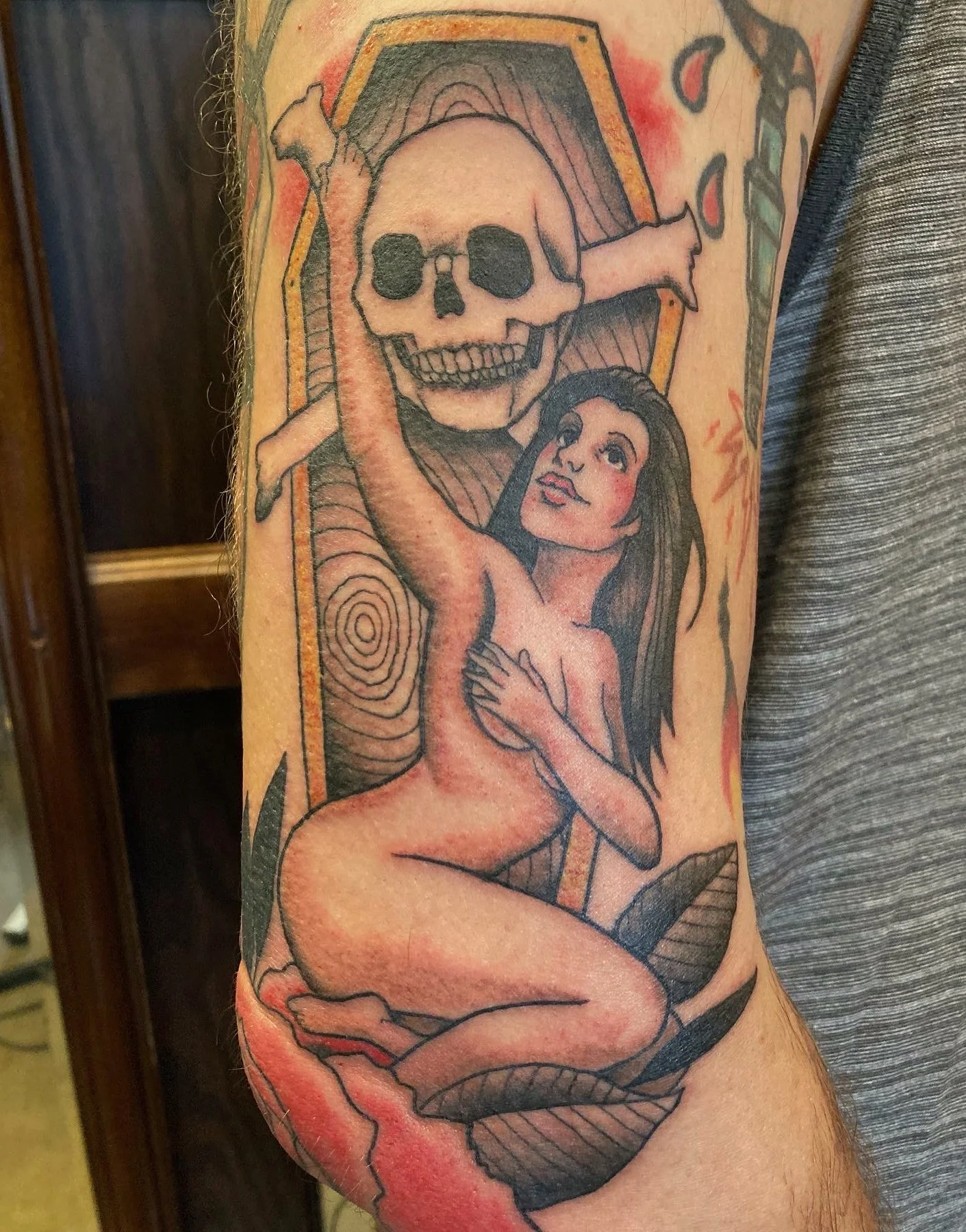 Tattoo of a woman holding a skull with a wooden coffin in the background and various plants around.