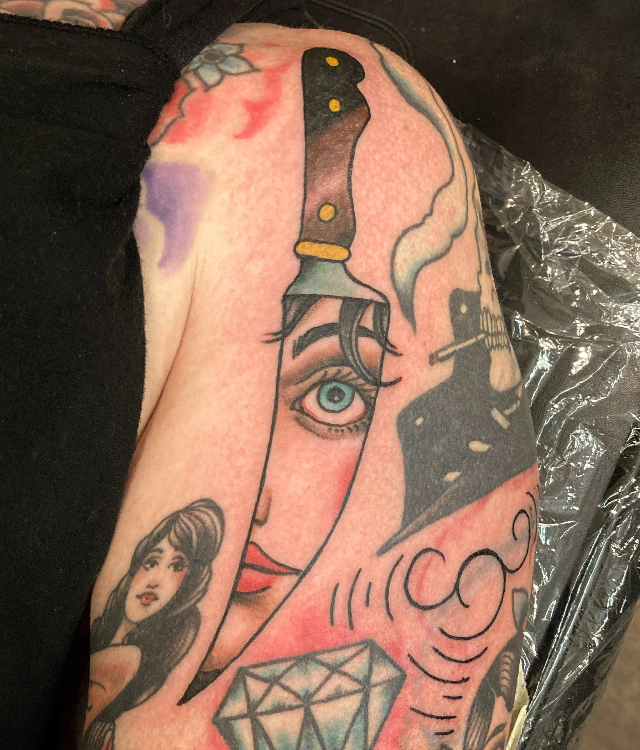 Tattoo of a dagger with an eye and a woman’s face, with additional small tattoos including a diamond and a woman’s portrait, on a person's arm.