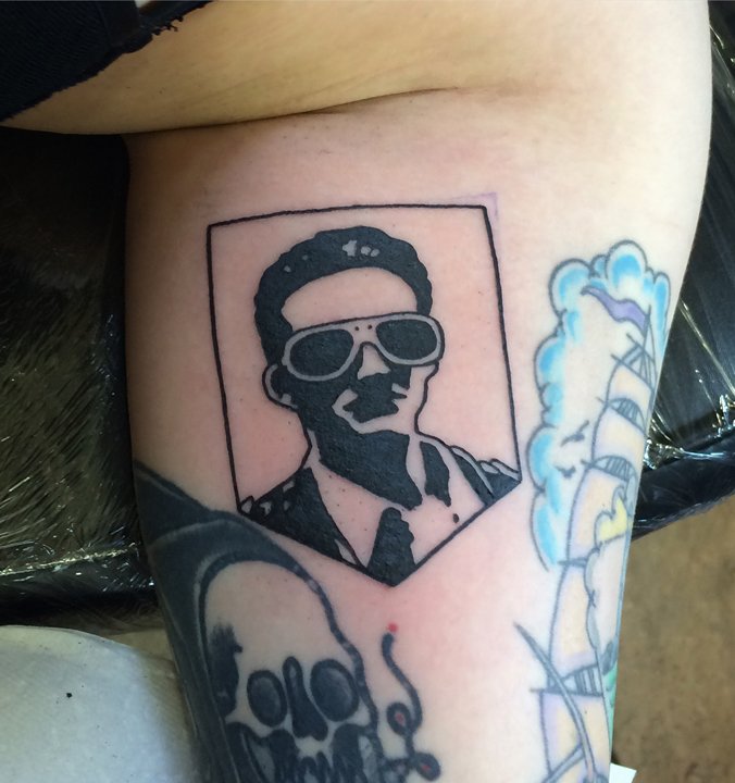 Tattoo of a man wearing sunglasses, a suit, and a tie in a framed portrait style, located on a person's arm.