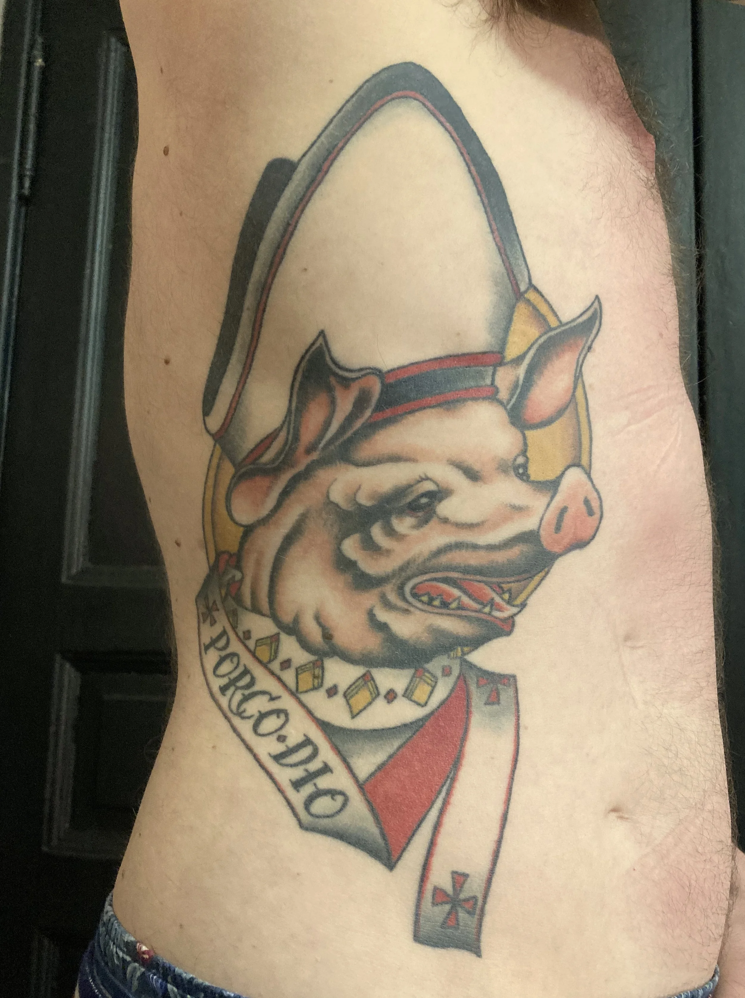 Tattoo of a pig's head with a baseball cap, bow, and jewelry, with a banner reading 'PORCO DIO'.