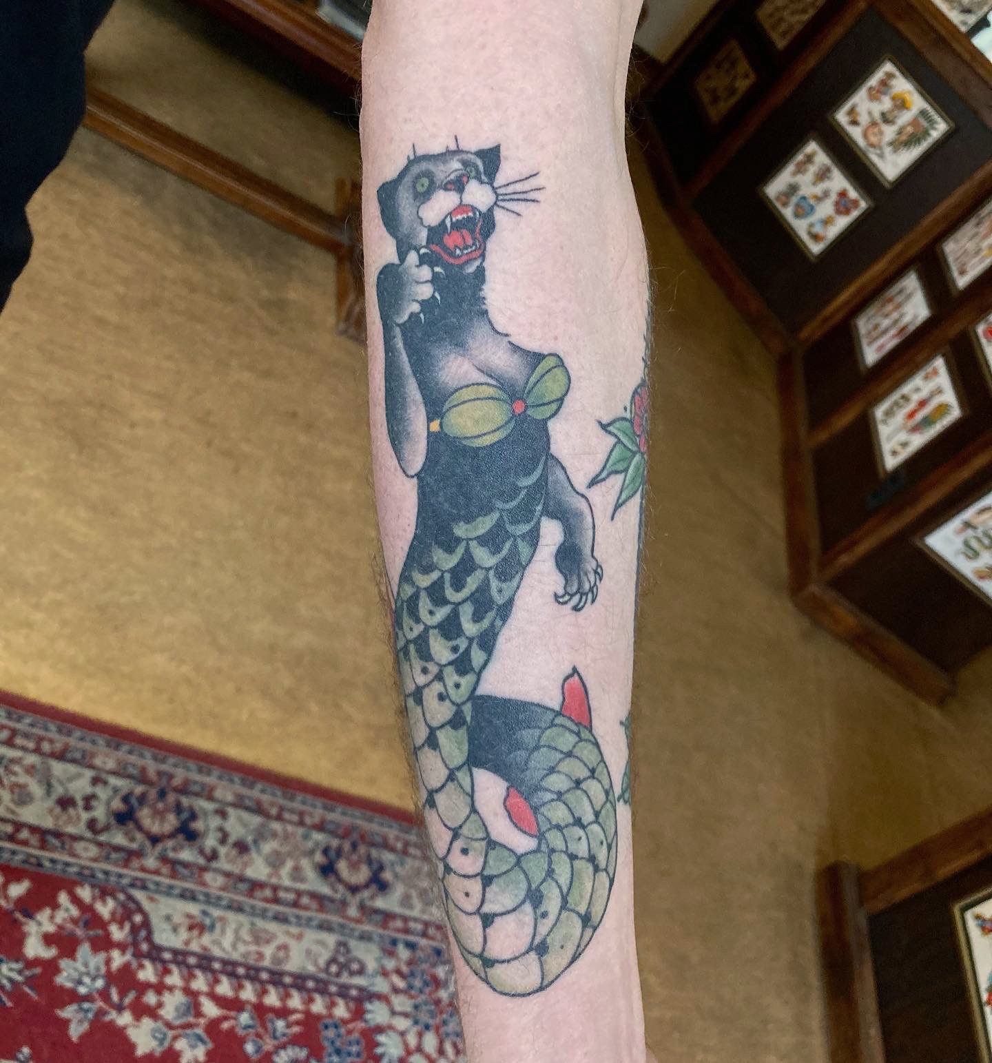 Tattoo of a black cat with a fish tail, holding a red flower.