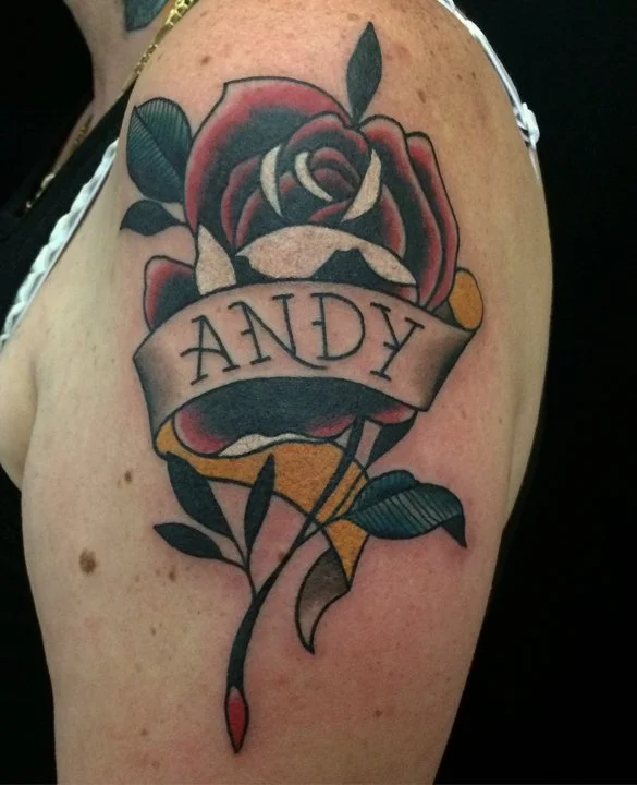 Tattoo of a blooming rose with green leaves, with a banner across it bearing the name 'Andy.'