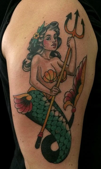 Tattoo of a mermaid holding a trident on an upper arm, featuring long dark hair with leaves, colorful tail with scales, and decorative fins.