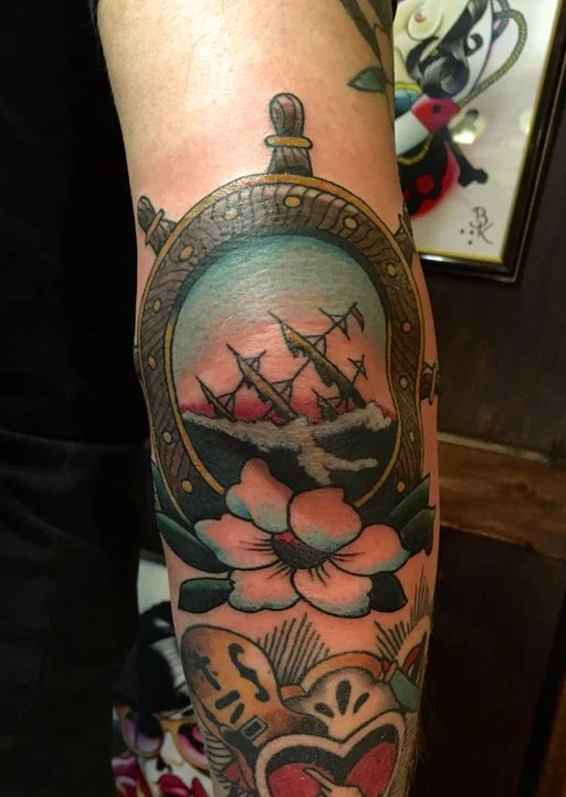 Tattoo of a vintage pocket watch with an illustration of three warships in battle, surrounded by a pink flower and green leaves.