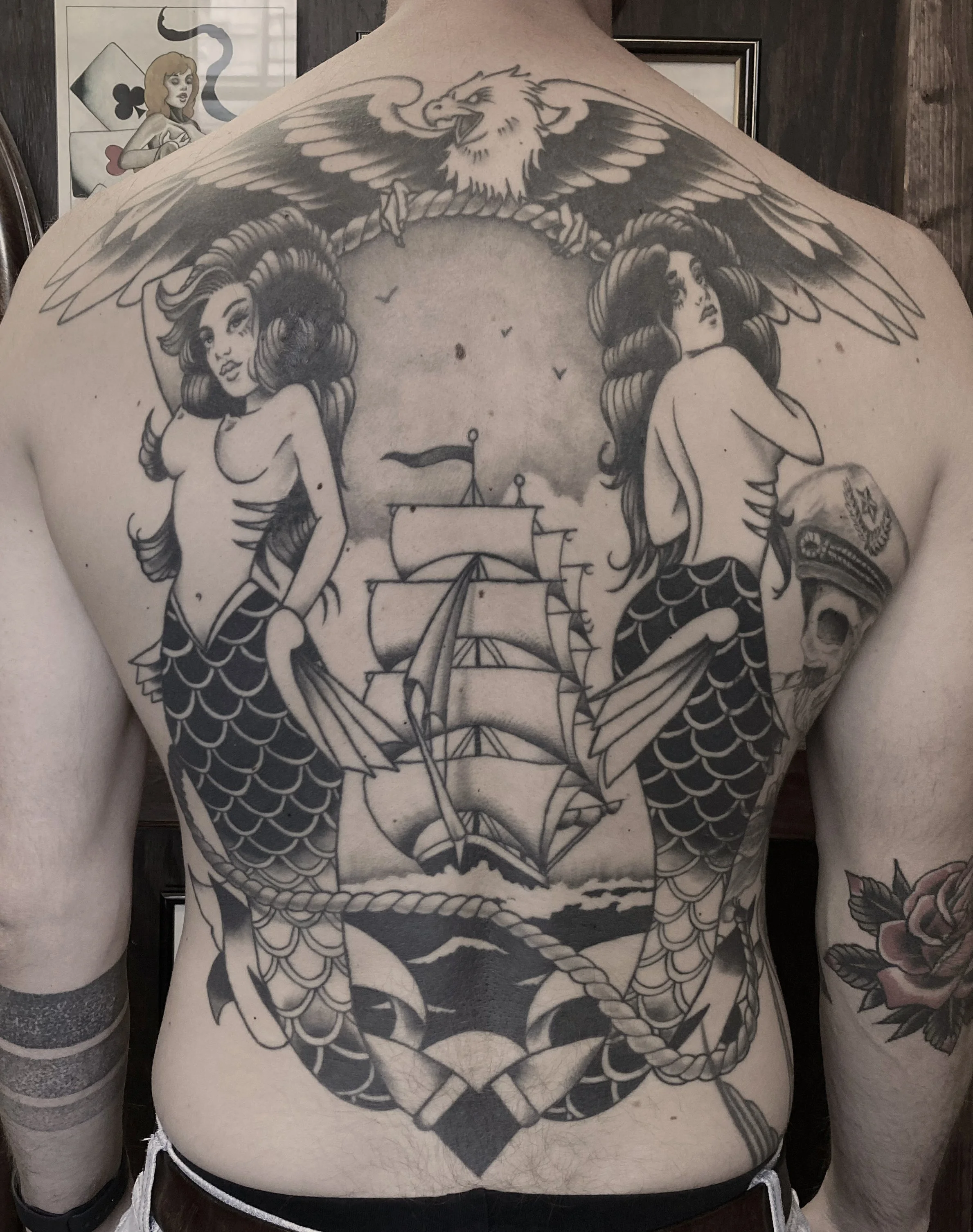 Black and gray tattoo on a person's back featuring two mermaids with long hair and scaly tails, a ship sailing on water, an eagle with spread wings.