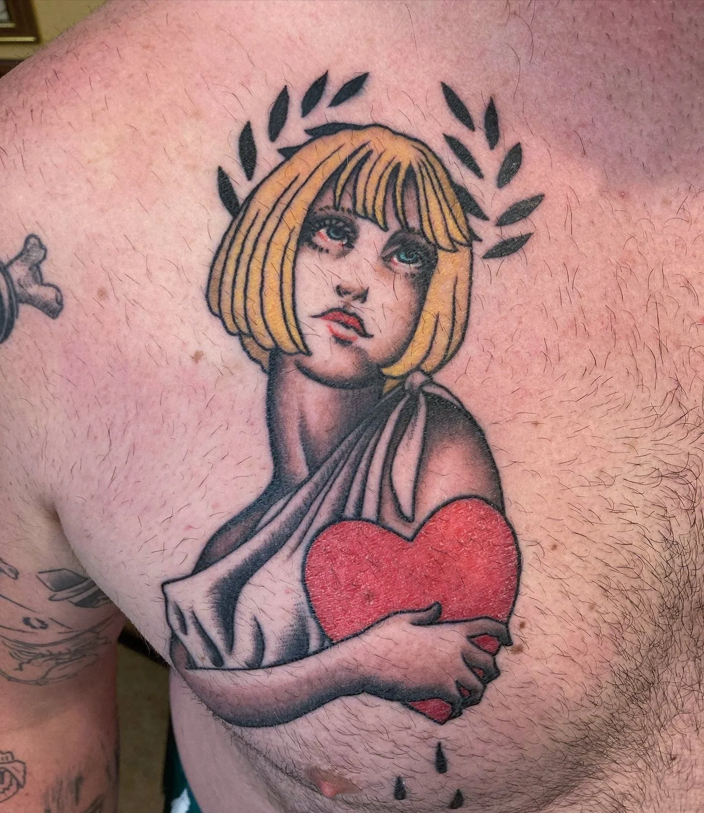 Tattoo of a woman with short blonde hair holding a red heart, surrounded by a laurel wreath.