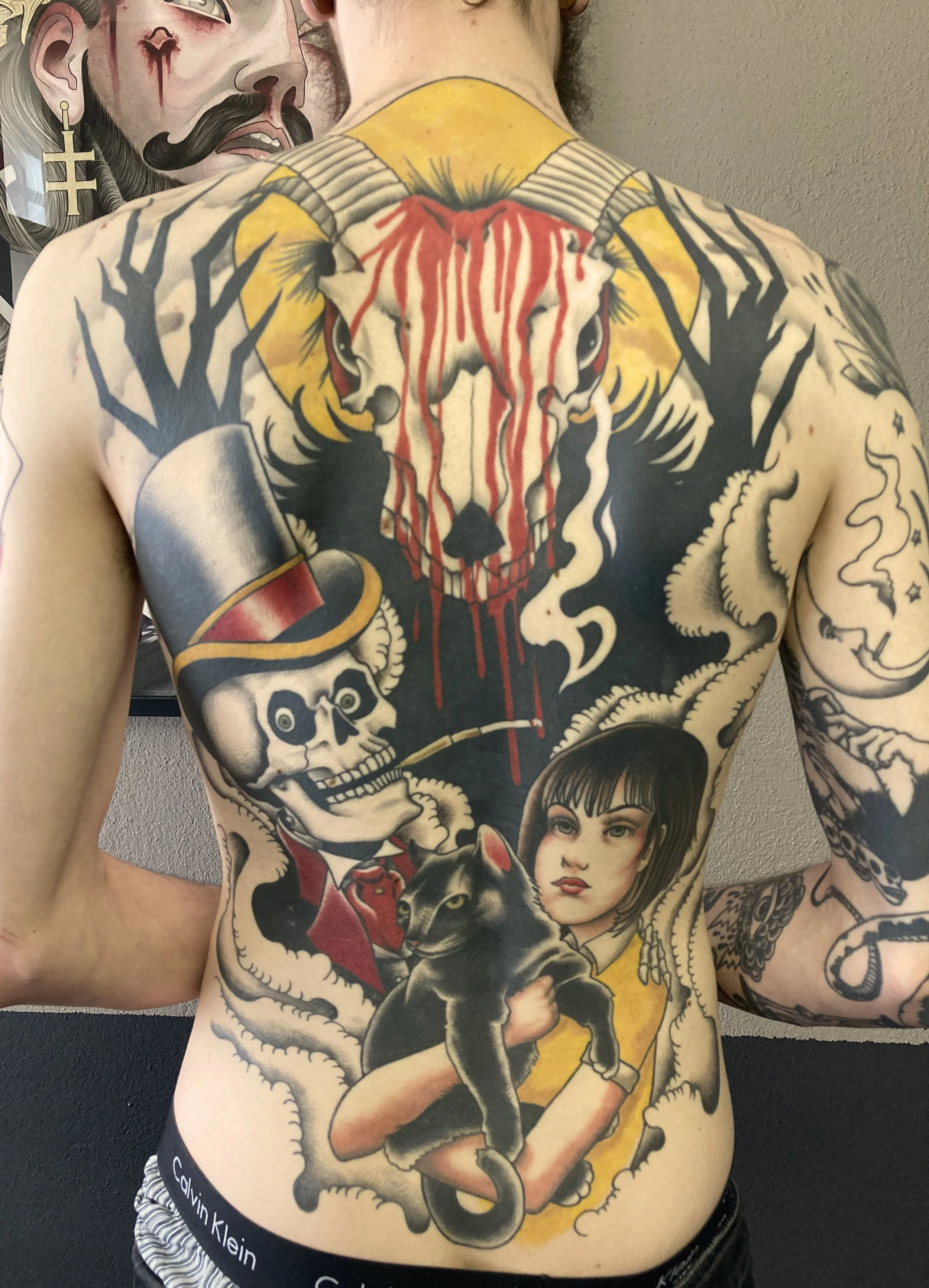 Colorful tattoo covering the person's chest and abdomen featuring a demons face with blood dripping, a skull wearing a red coat and top hat smoking a pipe, a black cat, a young girl with short hair, and various other design elements.