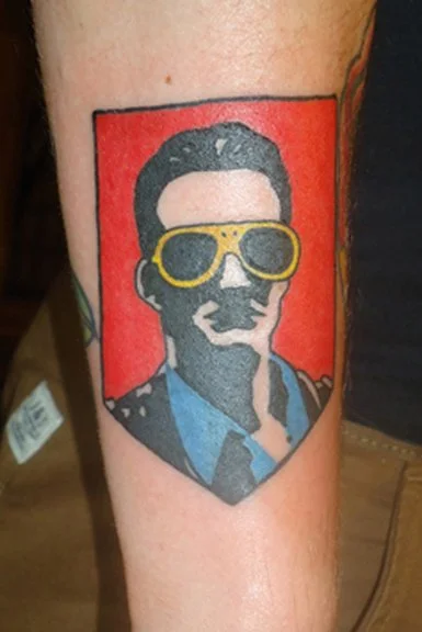 Tattoo of a man with dark hair, sunglasses, wearing a blue collared shirt and a dark jacket, on a red background.