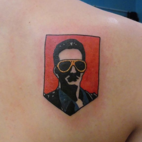 Tattoo of a man with sunglasses, and dark hair on the upper shoulder.