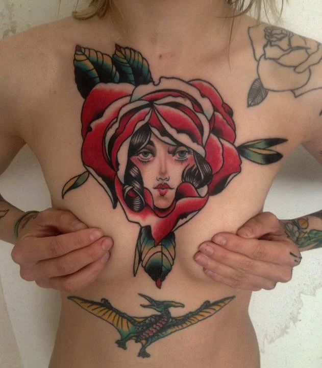 Tattoo of a woman's face with dark hair and pink cheeks, surrounded by a red rose and green leaves on the chest. Additional tattoos include a black and white rose on the shoulder and a dragon with outstretched wings below the chest.
