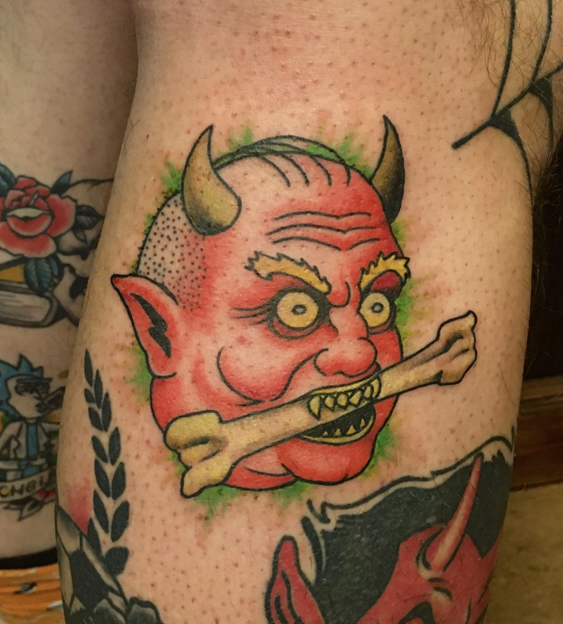 Tattoo of a cartoonish red devil with horns and yellow eyes, holding a bone in its mouth, surrounded by black and green ink details.