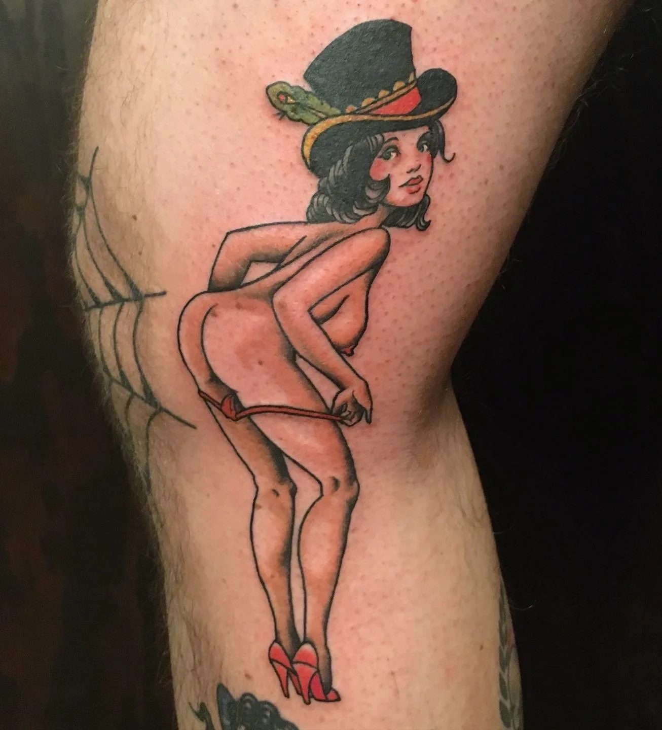 Tattoo of a vintage pin-up girl wearing a large hat, red high heels, and minimal clothing, sitting on a web.