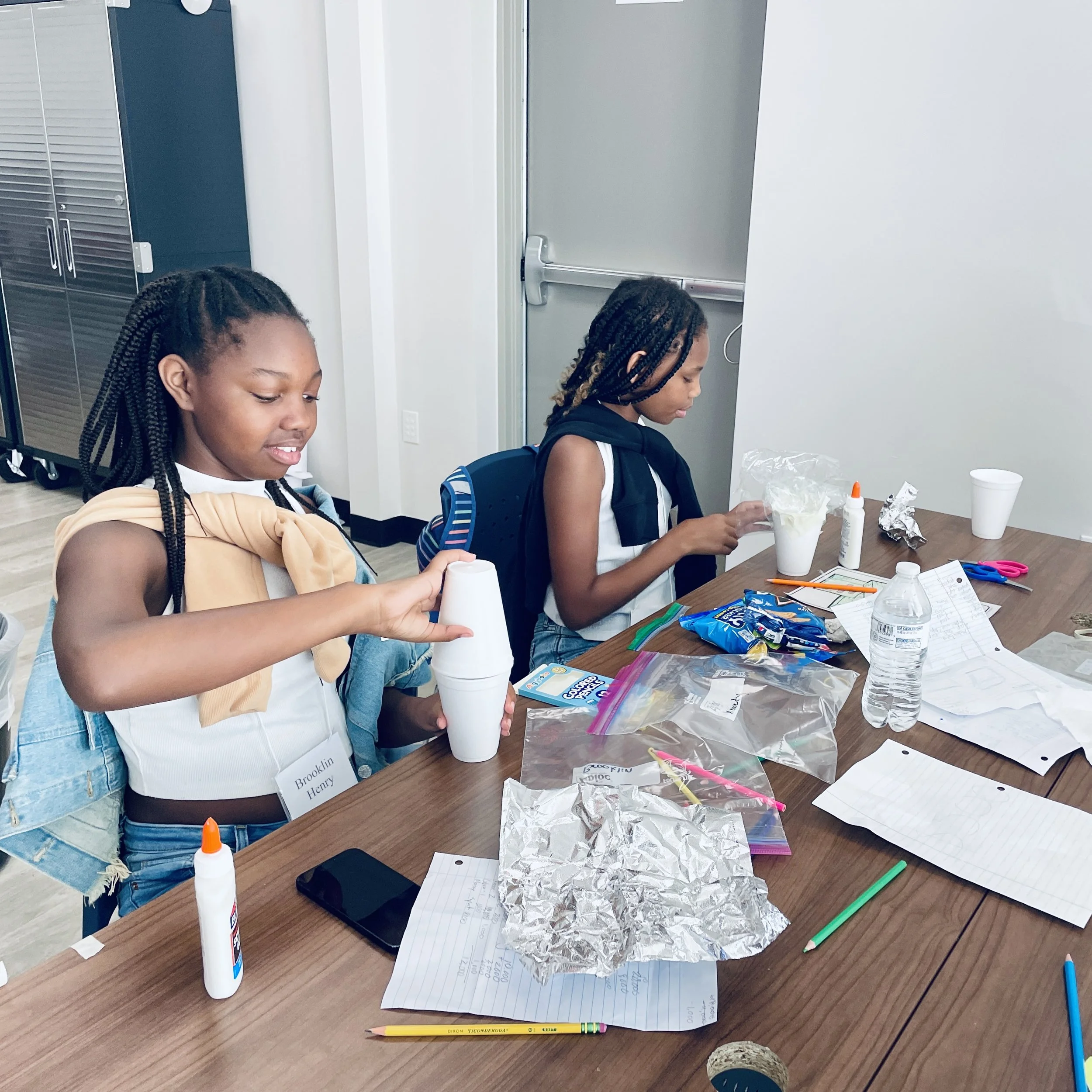 Two young girls sitting at a table with craft supplies, including glue, scissors, paper, and aluminum foil, engaging in arts and crafts activities.