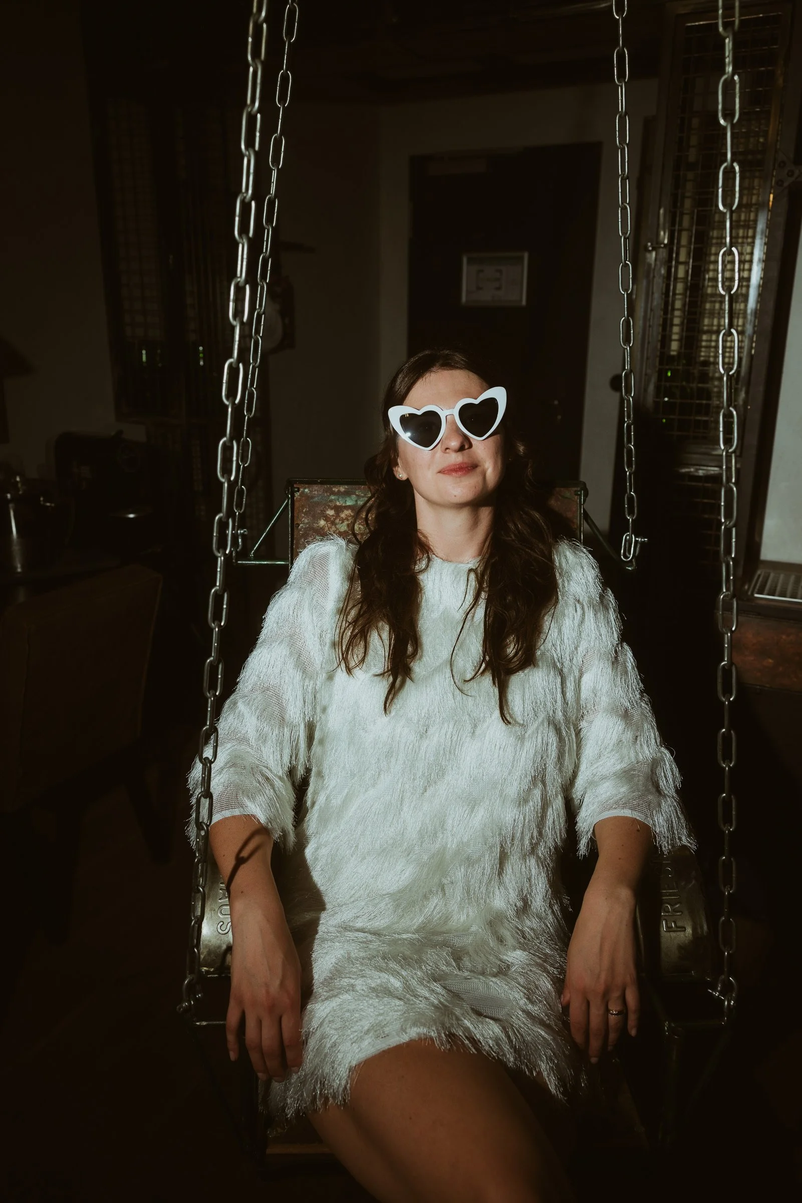 Eine Frau mit dunklen, lockigen Haaren trägt Herzförmige Sonnenbrille und sitzt auf einer Schaukel im dunklen Raum, gekleidet in einen weißen, federartigen Kleid.