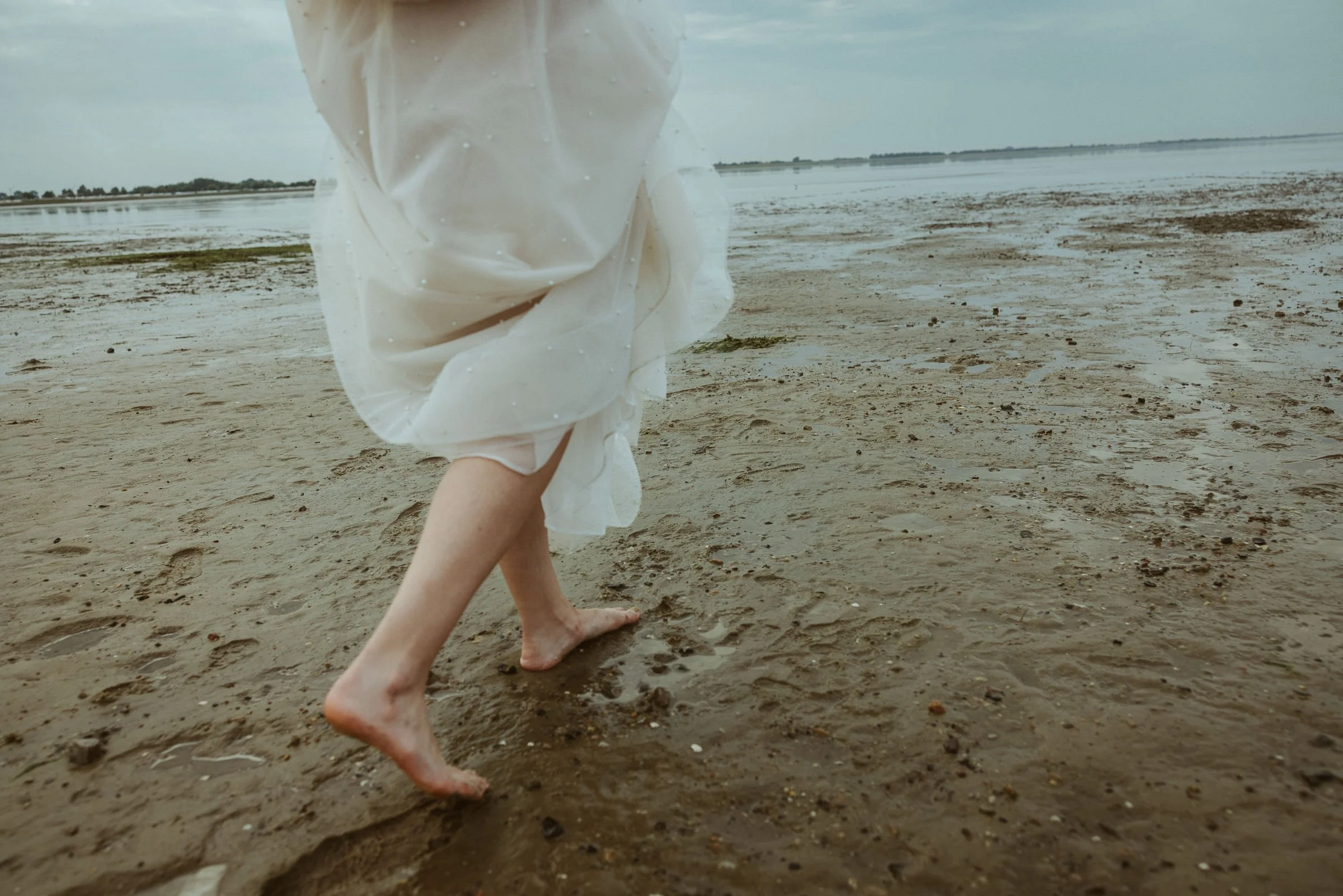 Person läuft barfuß am Strand entlang, hält ein weißes, nasses Tuch, im Hintergrund Wasser und Himmel.