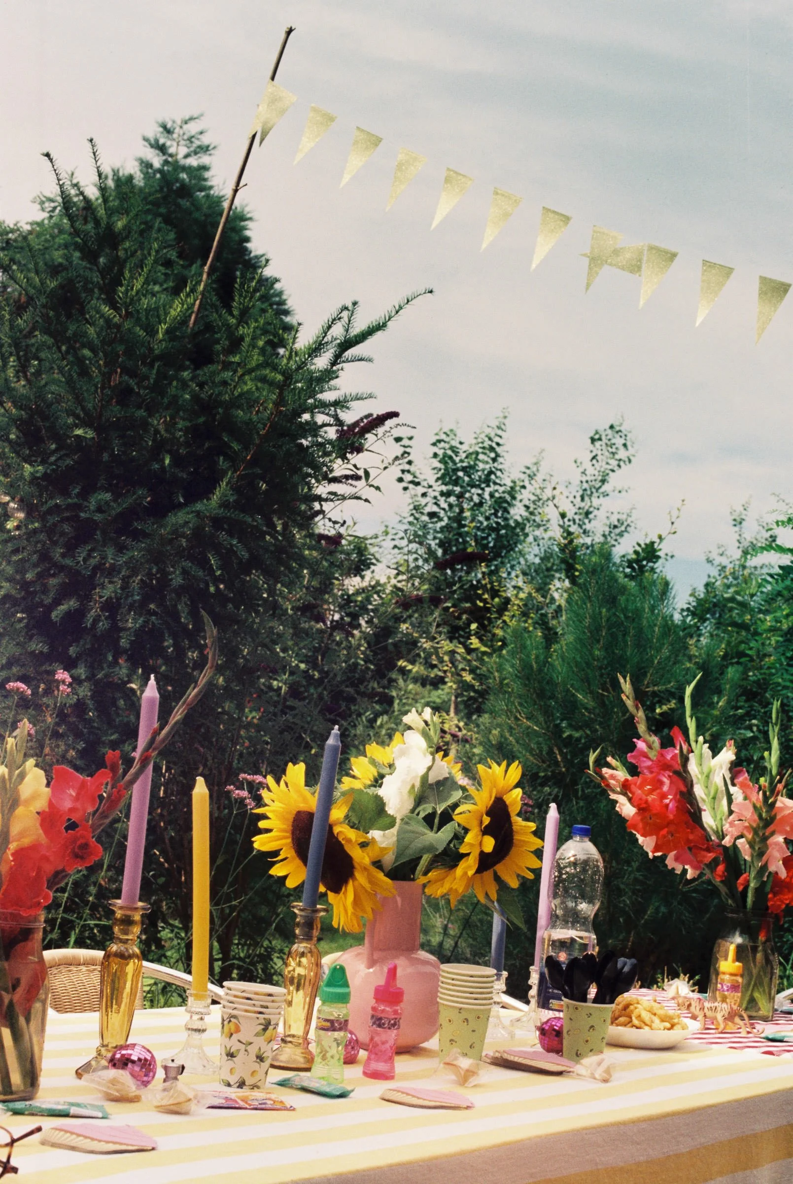 Ein Buffet mit bunten Blumen, Kerzen, Getränken und Snacks, im Freien bei bewölktem Himmel, bei einer Party oder Feier im Garten.