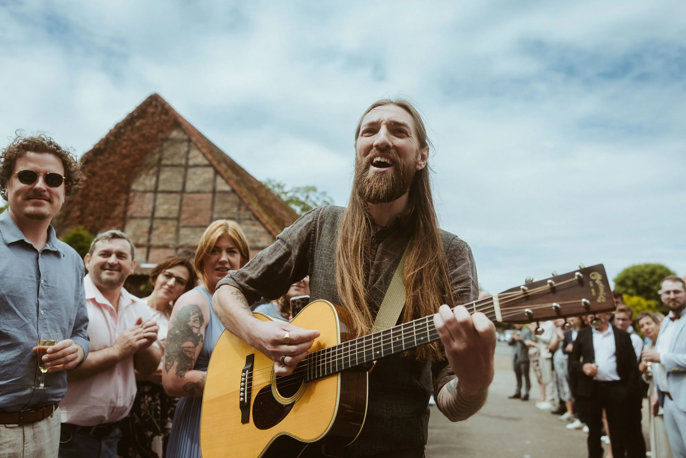 Ein Mann mit langem Haar und Bart spielt Gitarre auf einer Feier im Freien, umgeben von mehreren Menschen, mit historischem Fachwerkhaus im Hintergrund, bei bewölktem Himmel.
