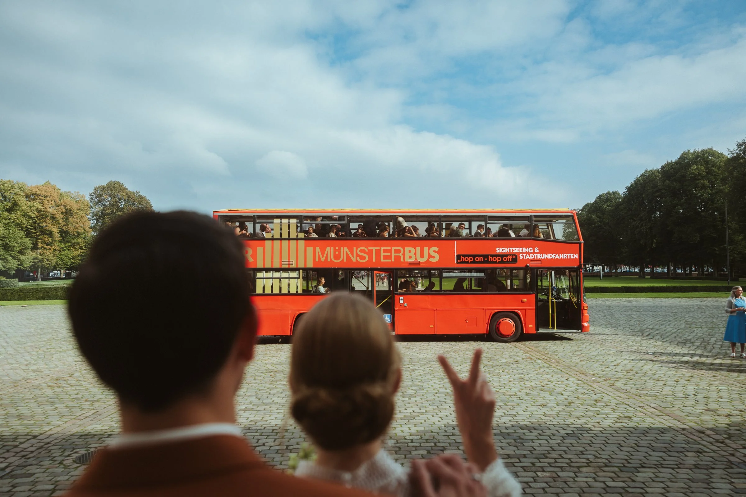 Ein roter Doppeldecker-Bus mit der Aufschrift 'Münsterbus' fährt durch einen Park, während Menschen auf den Bus und auf den Betrachter zugreifen, die den Bus beobachten.