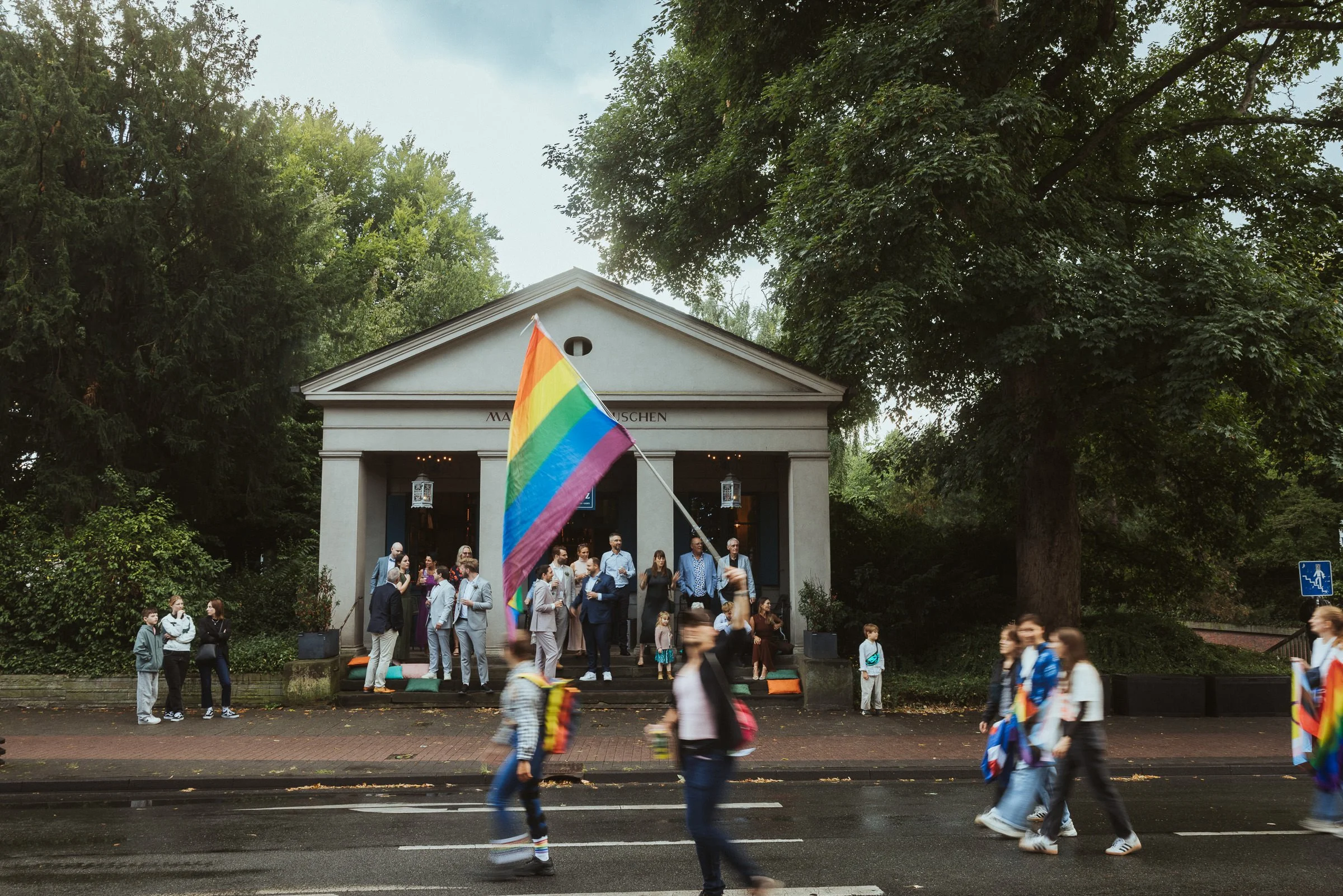 Menschen versammeln sich vor einem weißen Gebäude mit Säulen, während eine Person eine Regenbogenfahne schwenkt, um die Pride-Parade oder eine LGBTQ+-Veranstaltung zu feiern. Im Hintergrund sind große Bäume und ein bewölkter Himmel sichtbar.