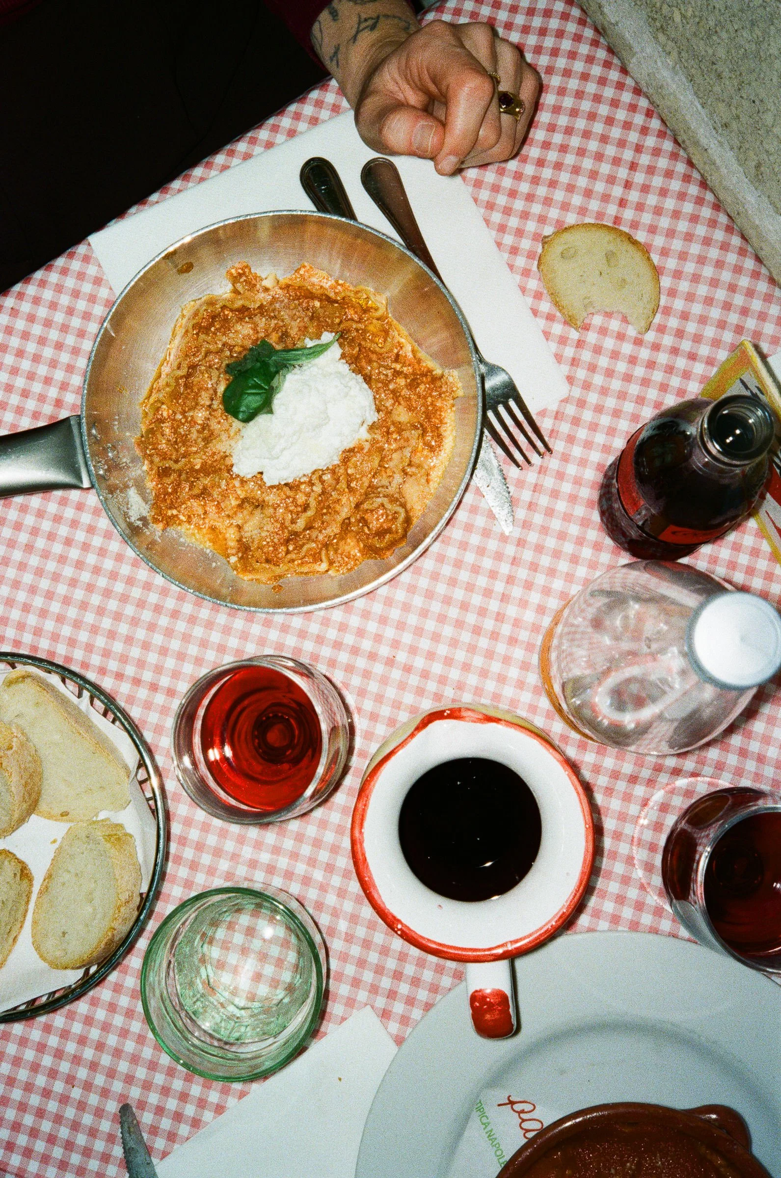 Ein Esstisch mit italienischer Mahlzeit, inklusive Schüssel mit Spaghetti und Tomatensoße, Brot, Gläser mit Rotwein, Wasser und dunkler Soße, auf rotes und weiß kariertes Tischtuch.
