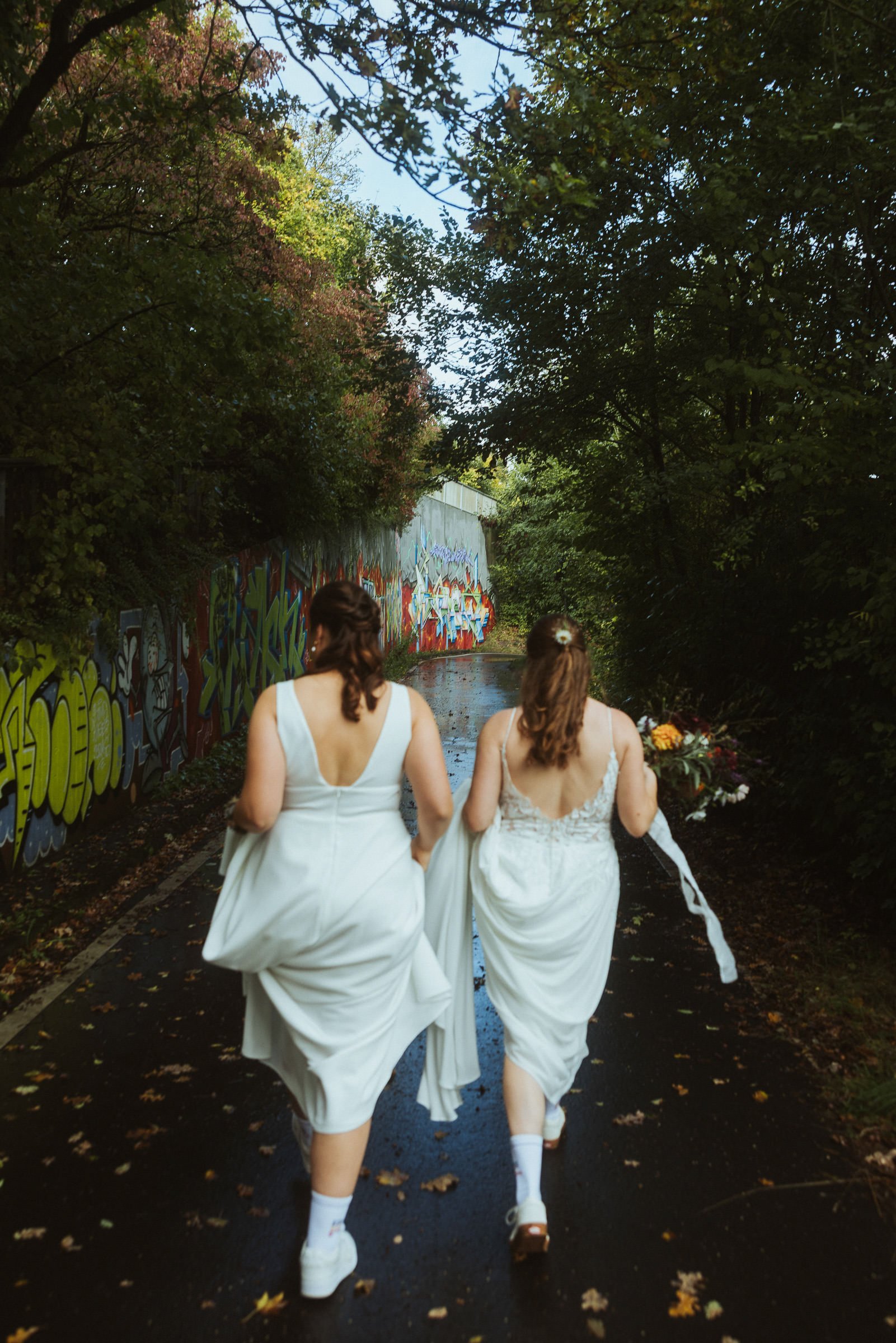 Drei Frauen laufen auf einem mit Laub bedeckten Weg, alle in weißen Kleidern, mit Blumen im Haar, auf dem Weg sind Graffiti an der Wand zu sehen, und es ist ein bewaldeter Bereich auf beiden Seiten.