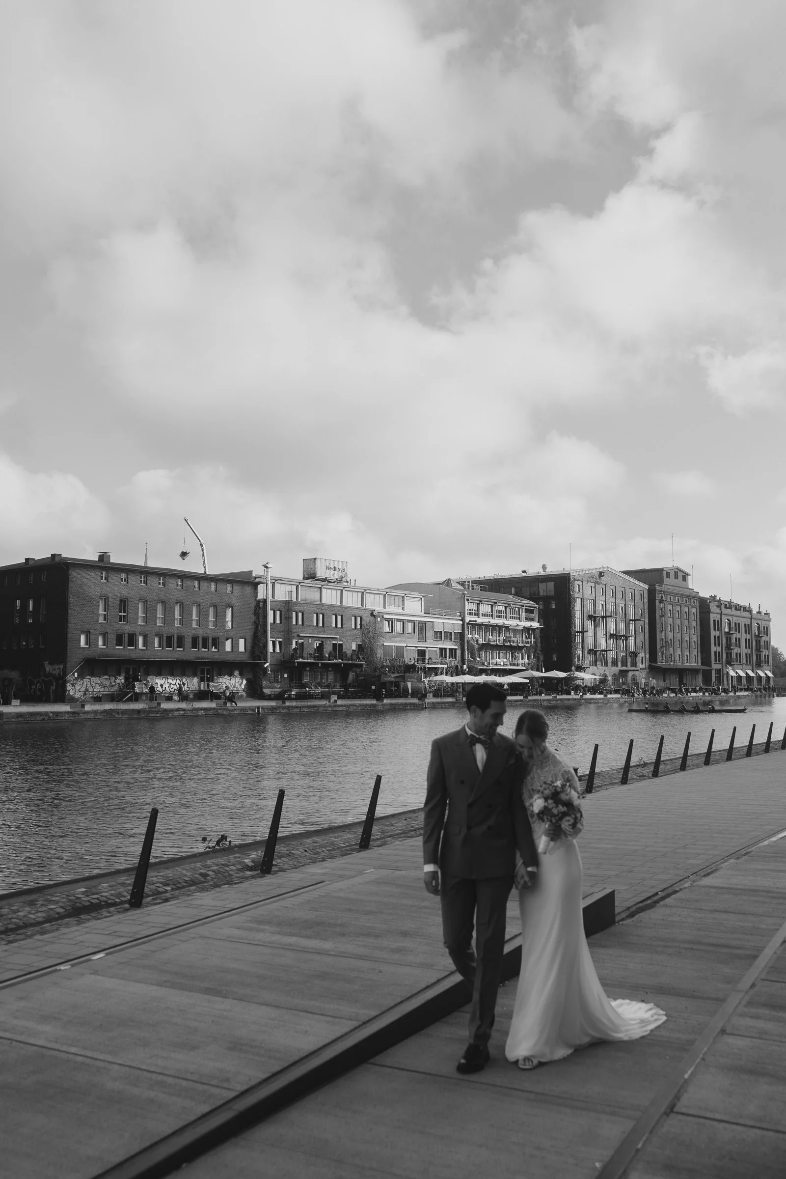 Ein Brautpaar läuft am Wasser entlang, im Hintergrund sind mehrstöckige Gebäude und Wolken am Himmel in Berlin.