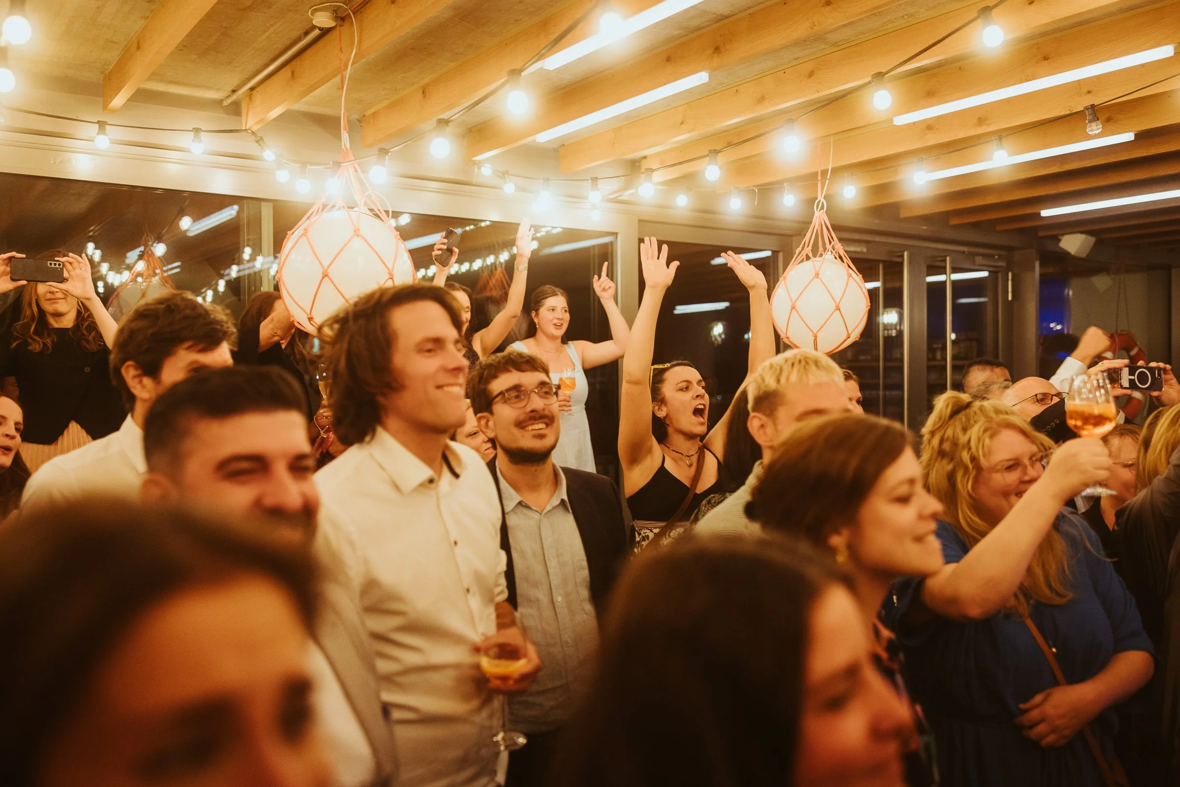 Menschen feiern in einem Raum mit Holzdecke und warmen Lichteffekten, einige heben die Arme, andere nehmen Fotos auf, während sie lachen und einen Drink halten.