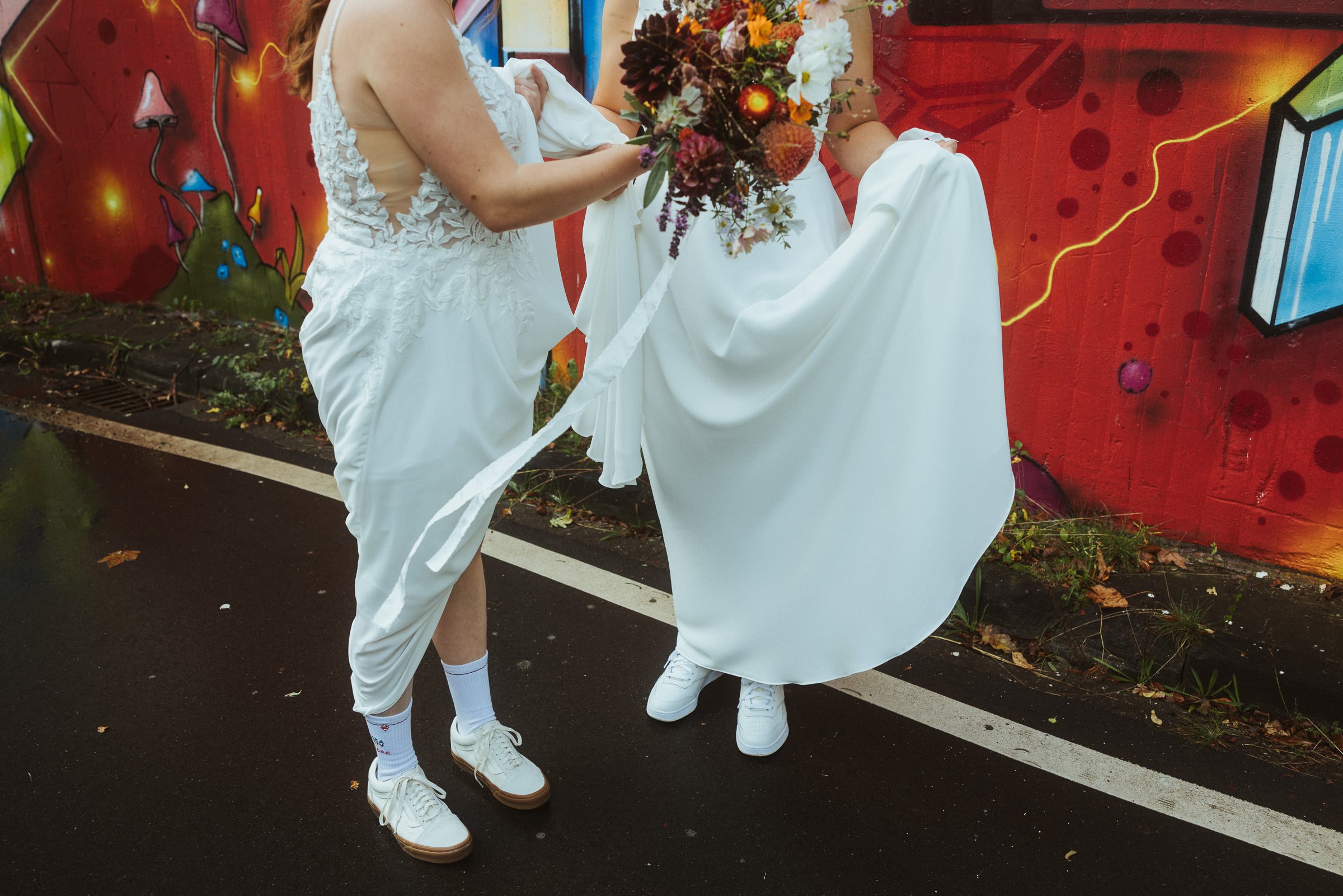 Zwei Menschen in weißen Hochzeitskleidern stehen nebeneinander auf einer Straße, eine hält einen Blumenstrauß. Sie tragen weiße Turnschuhe. Im Hintergrund eine bunte, graffitiartige Wand, die mit roten und anderen leuchtenden Farben bemalt ist.
