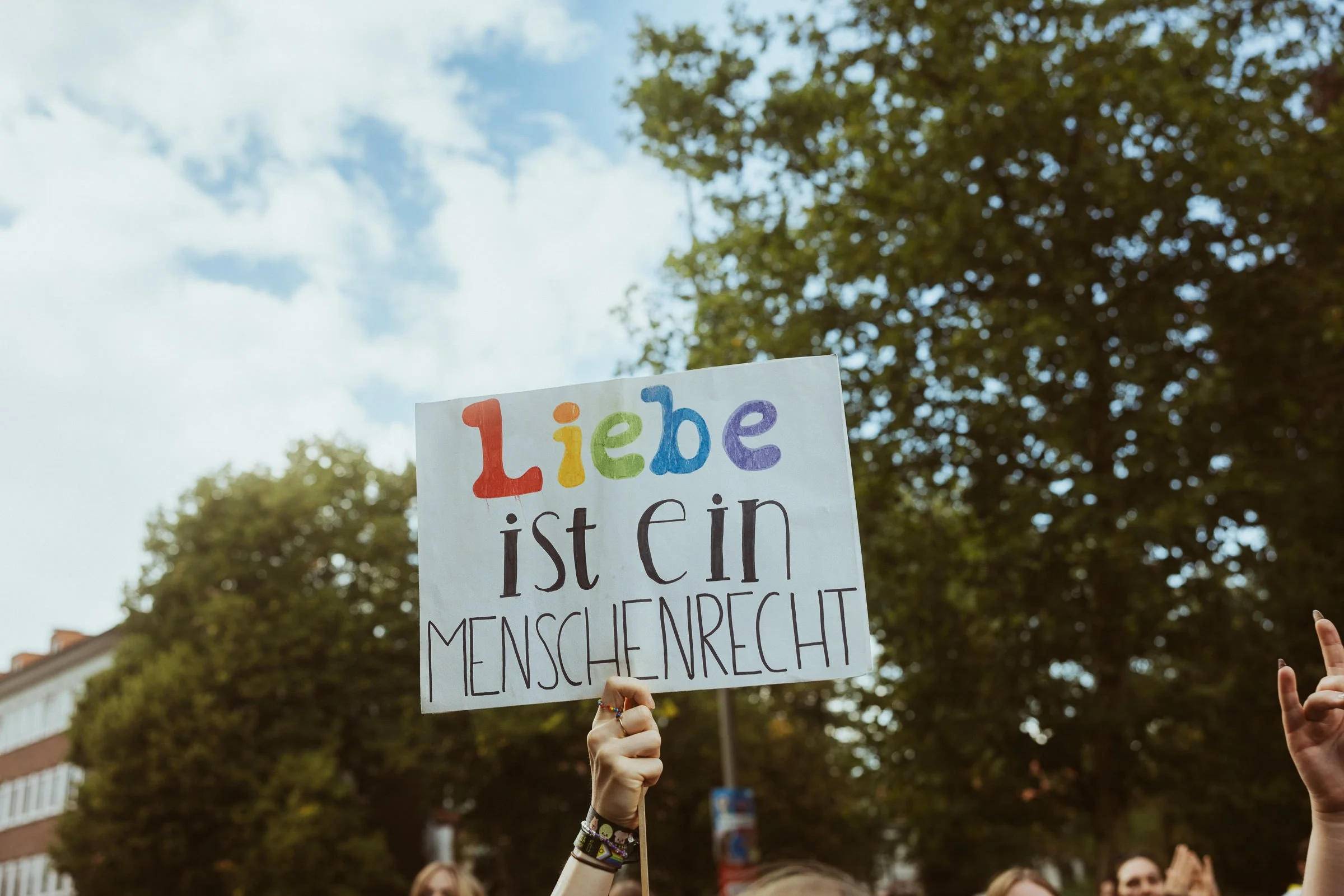 Menschen halten Protestplakat mit bunten Buchstaben, das sagt: Liebe ist ein Menschenrecht, bei einem Demonstrationszug im Freien an einem sonnigen Tag, im Hintergrund Bäume und Gebäude.