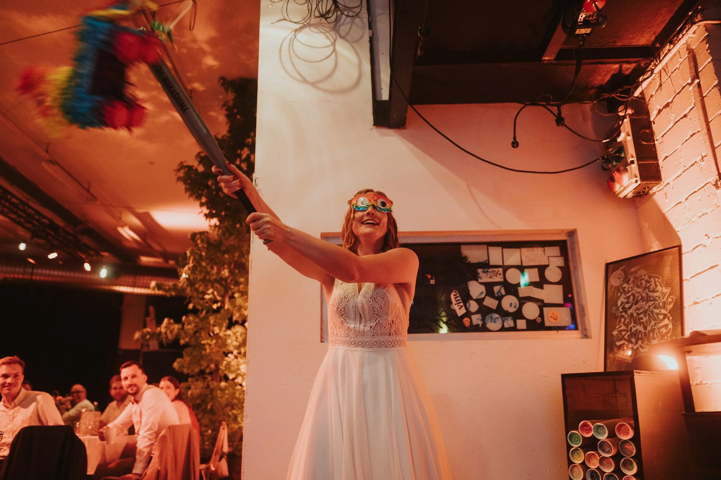 Eine lachende Frau mit buntem Partybrille tanzt mit einem Besen bei einer Party, während Gäste im Hintergrund sitzen und beobachten. Innenraum mit Dekorationen und einer Fensterwand.