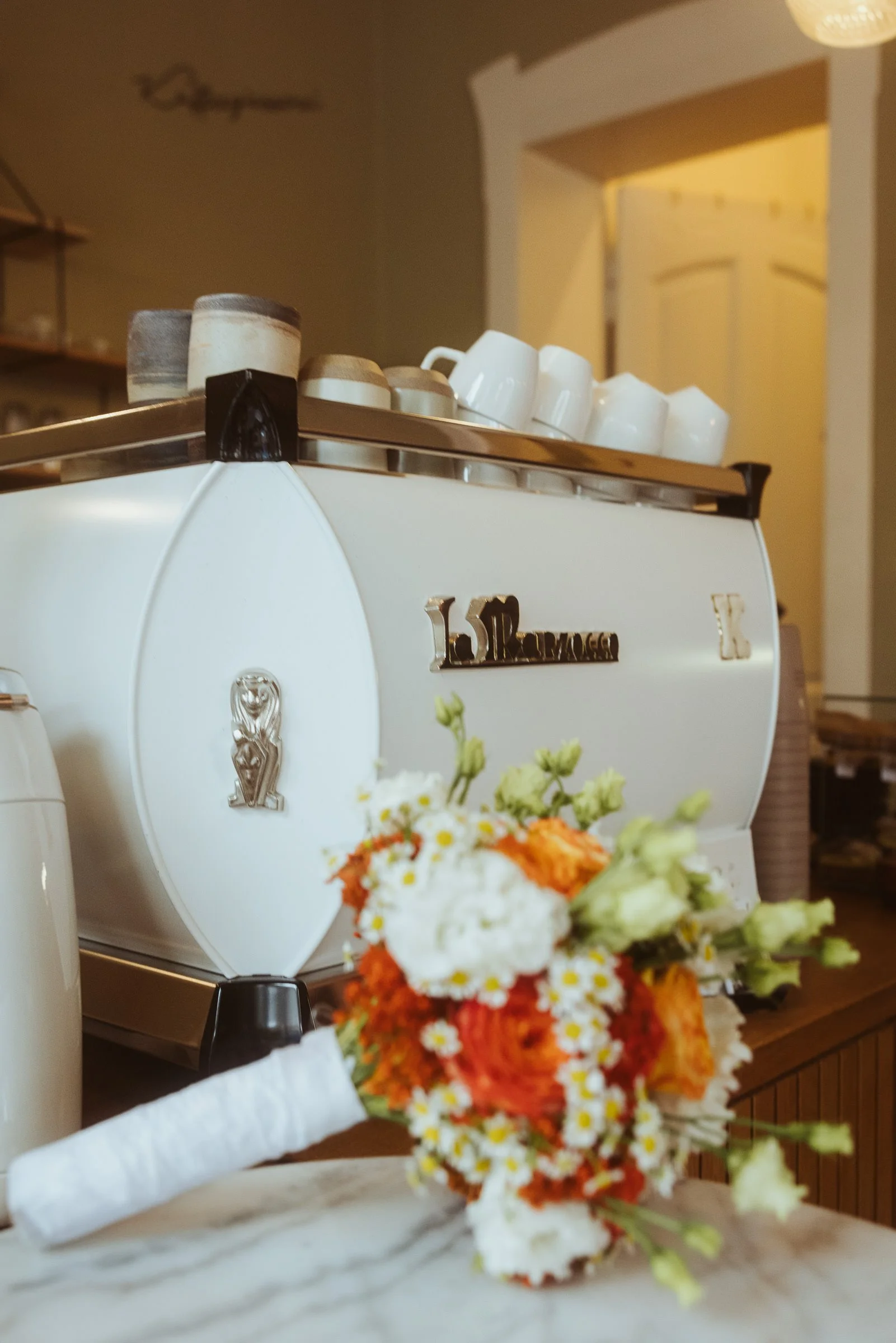 Blumenstrauß mit roten, weißen und gelben Blumen auf einer weißen Oberfläche, im Hintergrund eine Espressomaschine mit Tassen und Kaffeedosen auf einer Ablage.