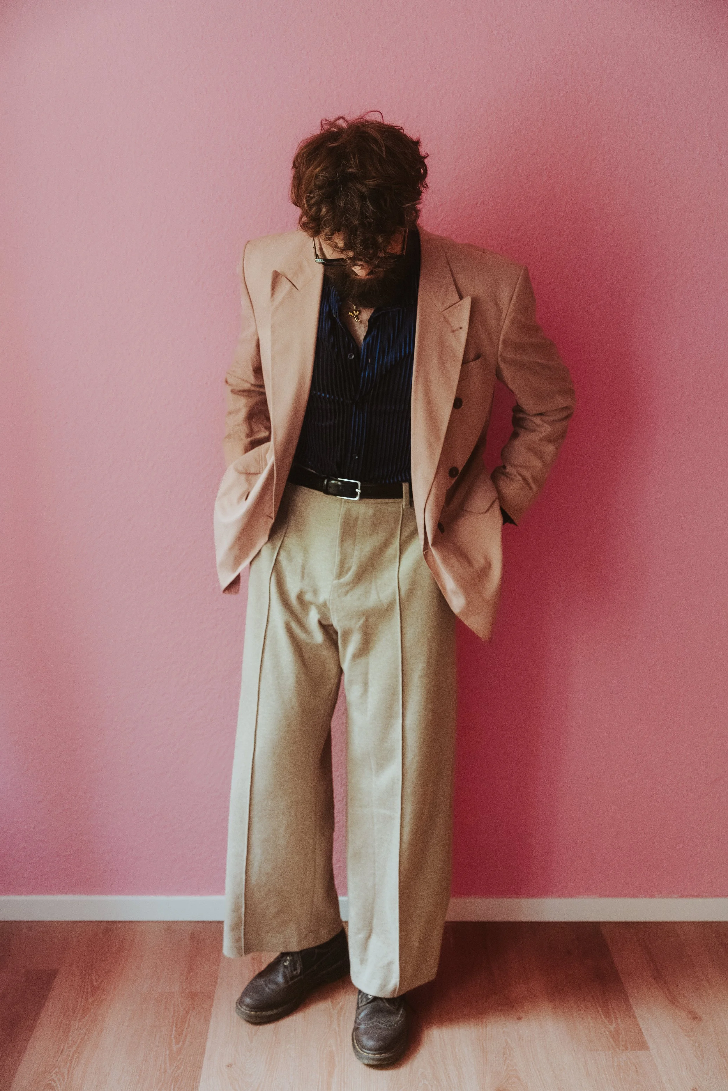 Mann mit dunklen, lockigen Haaren, Brille und Bart trägt beige Anzughose, schwarze Schuhe, schwarzen und blauen gestreiften Hemd und beige Blazer, steht vor pinker Wand.
