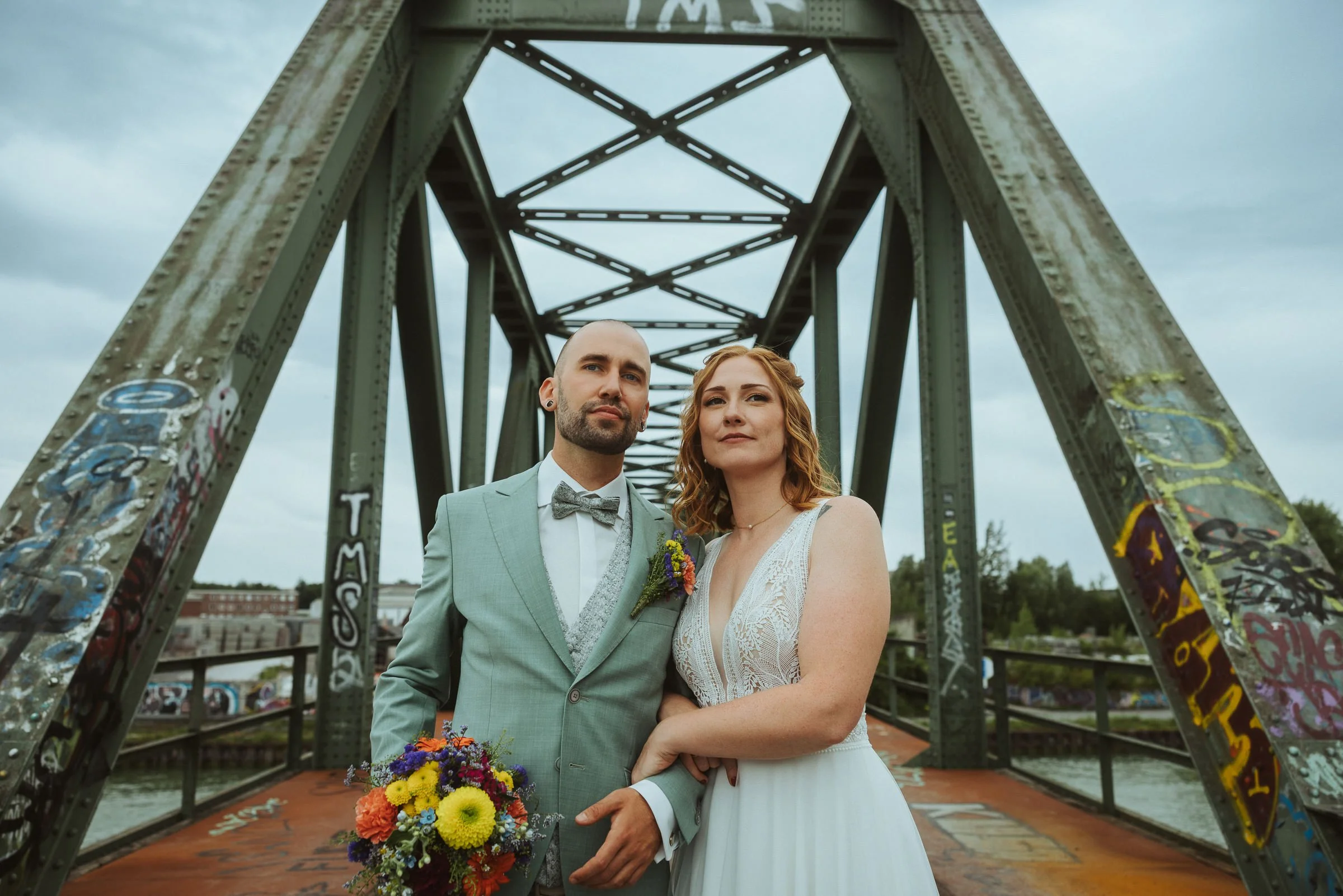 Ein frisch verheiratetes Paar posiert unter einer alten, graffiti-übersäten Eisenbrücke, der Mann trägt einen hellgrauen Anzug mit Fliegen, die Frau trägt ein weißes Hochzeitskleid und hält einen bunten Blumenstrauß.