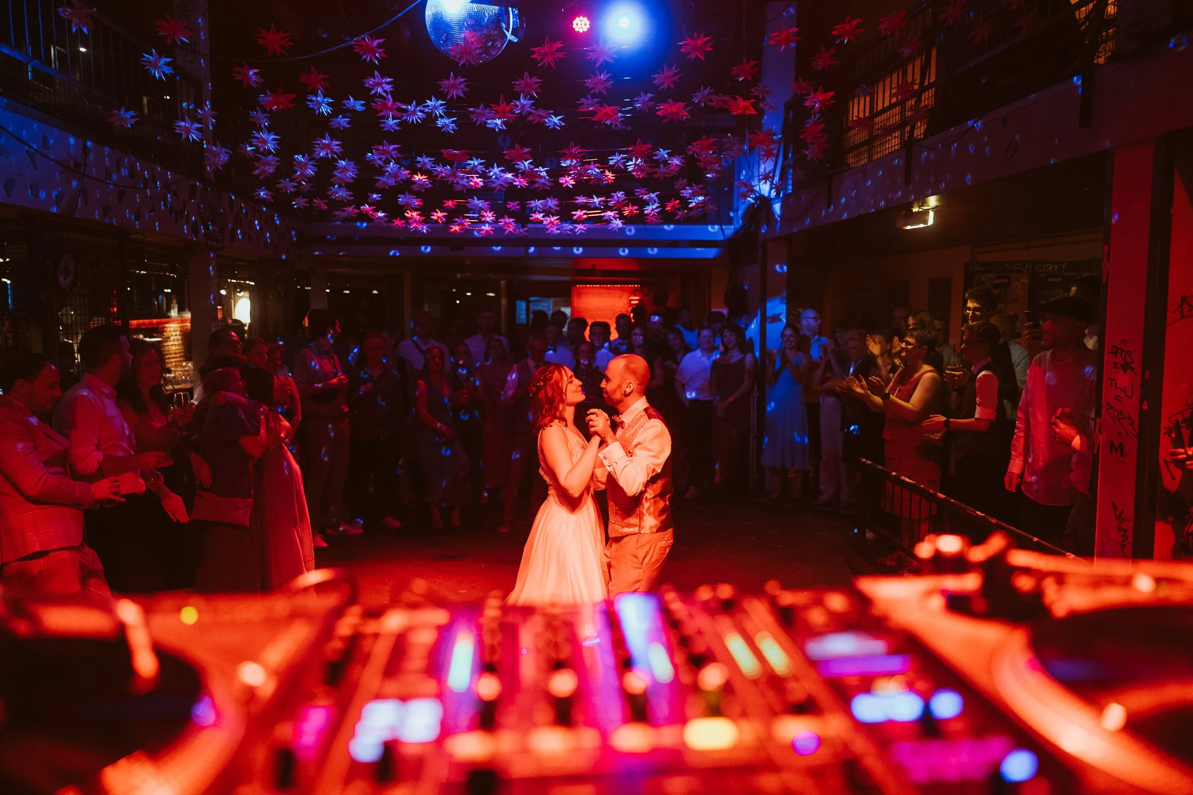 Paare tanzen in einer diskothekähnlichen Umgebung mit bunter Beleuchtung und Dekorationen, während einem DJ an einem Mischpult im Vordergrund zu sehen ist.