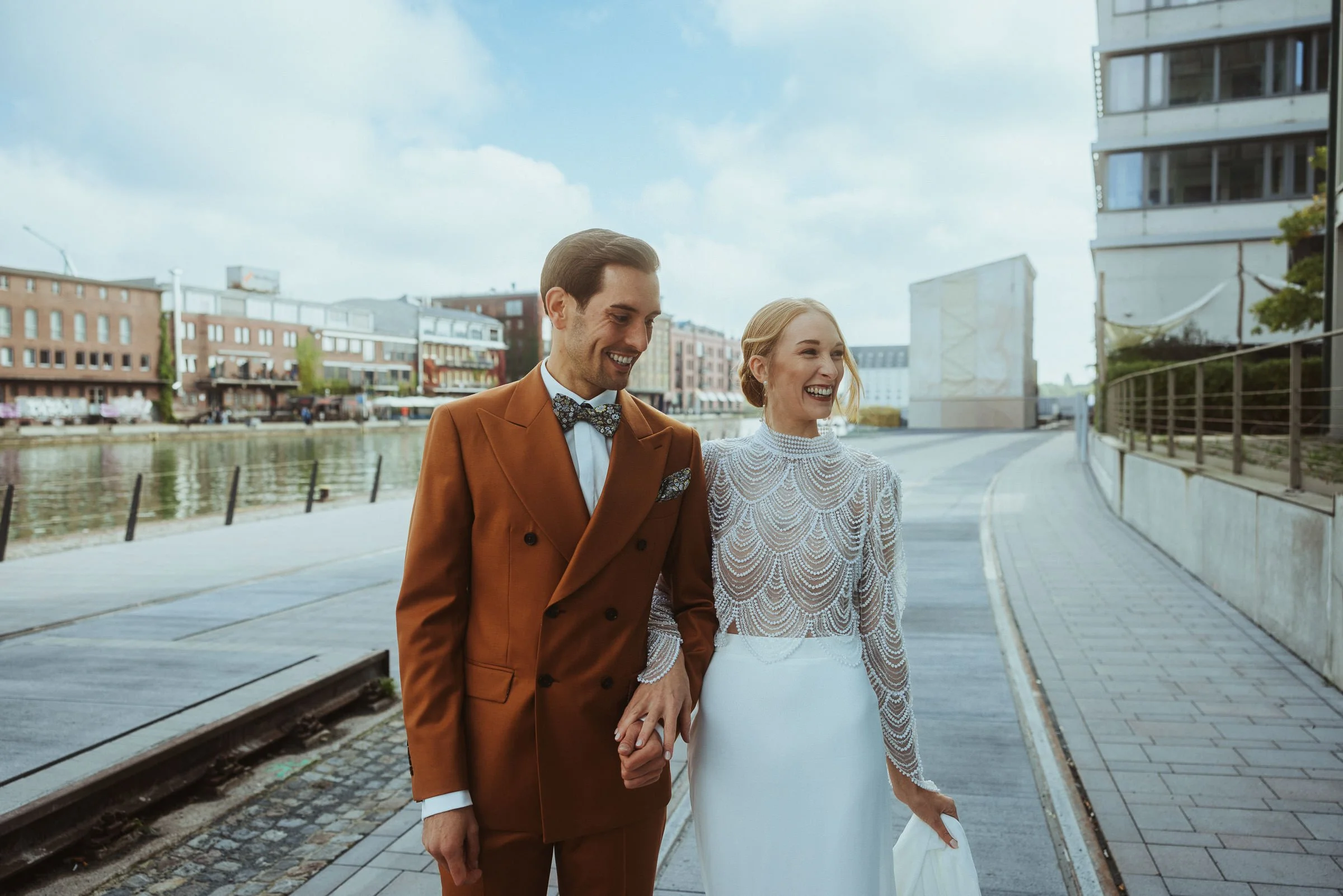 Ein Brautpaar, das lachend die Hand hält, bei einem Spaziergang entlang eines Flusses in einer Stadt mit modernen Gebäuden, während die Braut ein elegantes weißes Kleid trägt und der Bräutigam einen braunen Anzug mit Fliege. Es ist tagsüber mit wolki