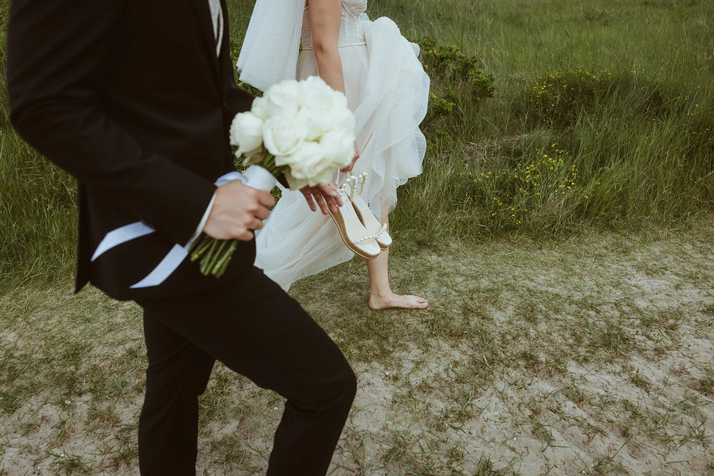 Ein Mann in einem schwarzen Anzug hält einen Blumenstrauß aus weißen Rosen, während eine Braut im weißen Hochzeitskleid barfuß im Gras steht, die Schuhe in der Hand.