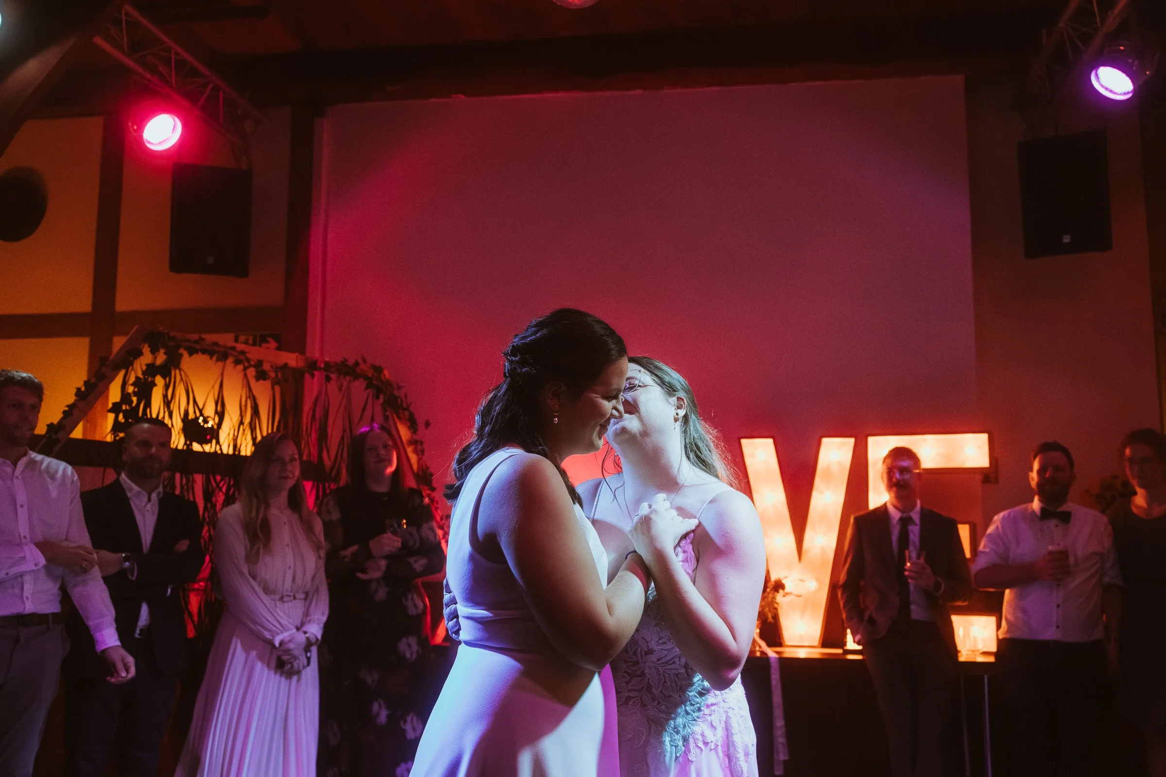 Zwei Frauen in einem Hochzeitskleid tanzen sich im Mittelpunkt bei einer Feier, umgeben von Gästen in einem festlich dekorierten Raum mit warmem Licht und großen Buchstaben, die für die Hochzeitsfeier stehen.
