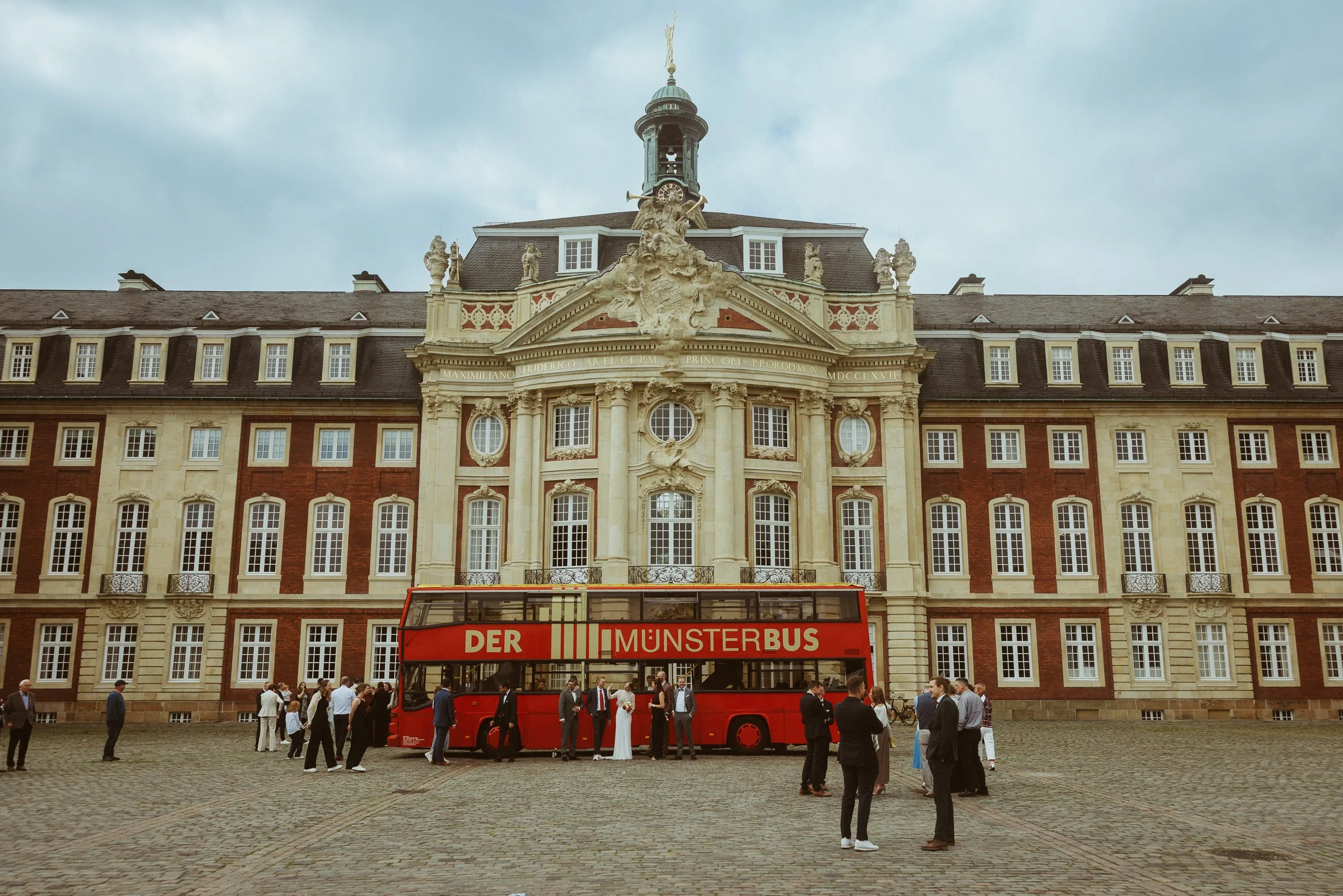 Ein roter Doppeldeckerbus mit der Aufschrift "Der MUNSTERBUS" steht vor einem historischen Gebäude mit barocker Architektur, das eine Statue und ein Uhrturm auf dem Dach hat. Es sind mehrere Menschen im öffentlichen Platz unterwegs.