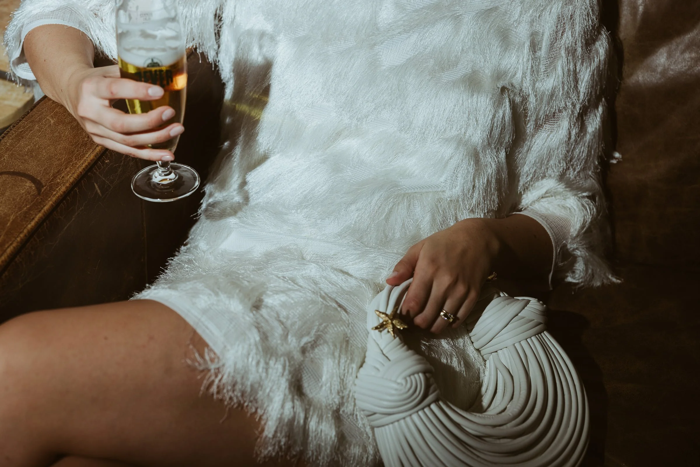 Person hält ein Glas Bier in der Hand, trägt ein weißes Outfit mit Perlen und trägt Ringe. Sitzt auf einem Holzstuhl, die Umgebung wirkt rustikal.