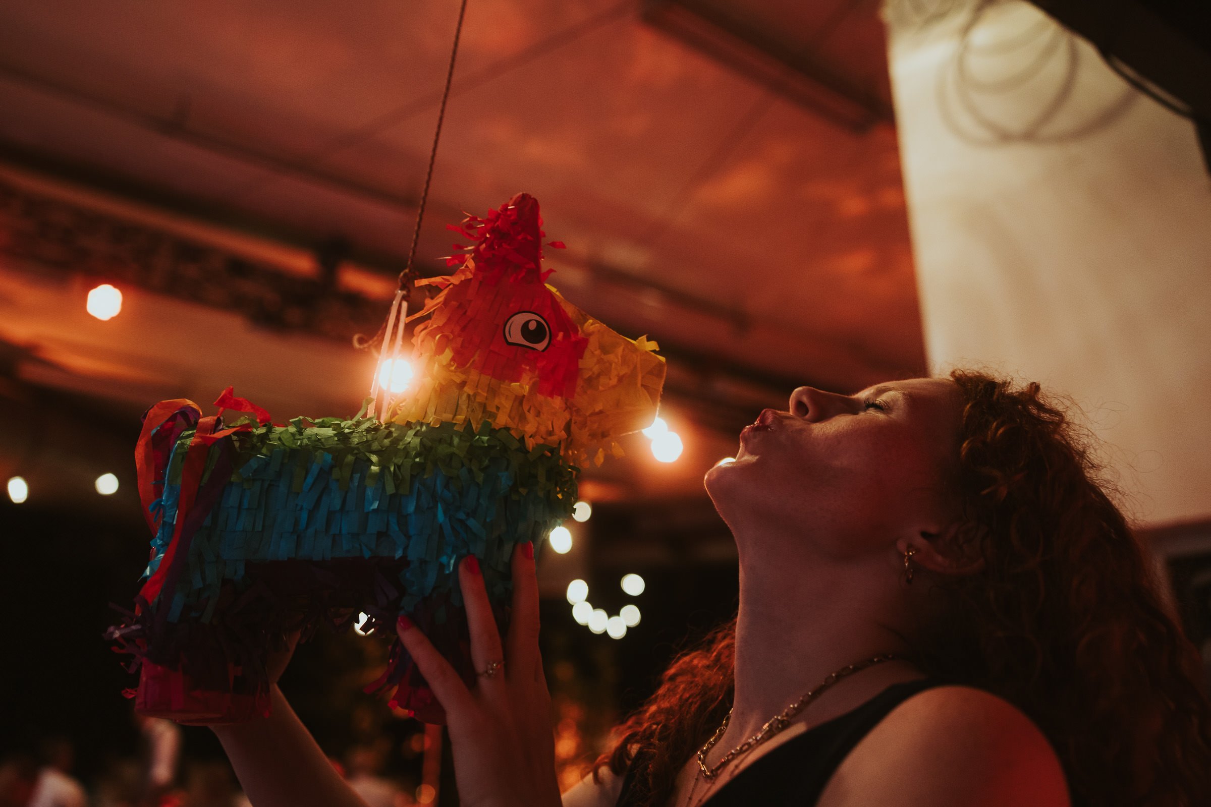 Junge Frau bei einer Fiesta mit einer bunten Piñata, die sie küsst, im Hintergrund hängen Lichter und es ist dunkel.