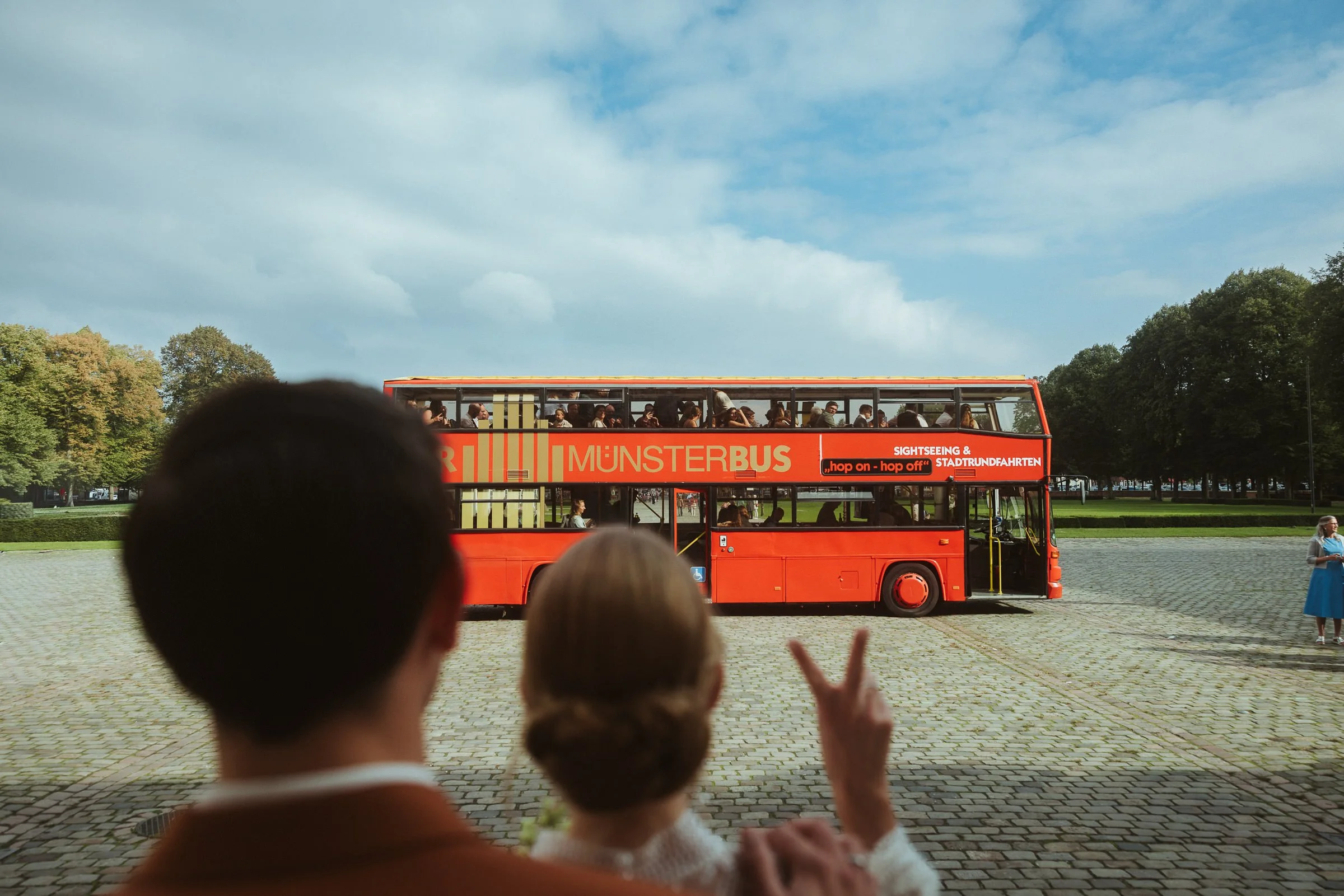 Ein roter Doppeldeckerbus mit der Aufschrift "Münster Bus" und "Sightseeing & Stadtrundfahrten" fährt durch einen Park. Es sitzen viele Touristen im Oberdeck des Busses. Im Vordergrund sind Kinder, die den Bus beobachten, zu sehen.