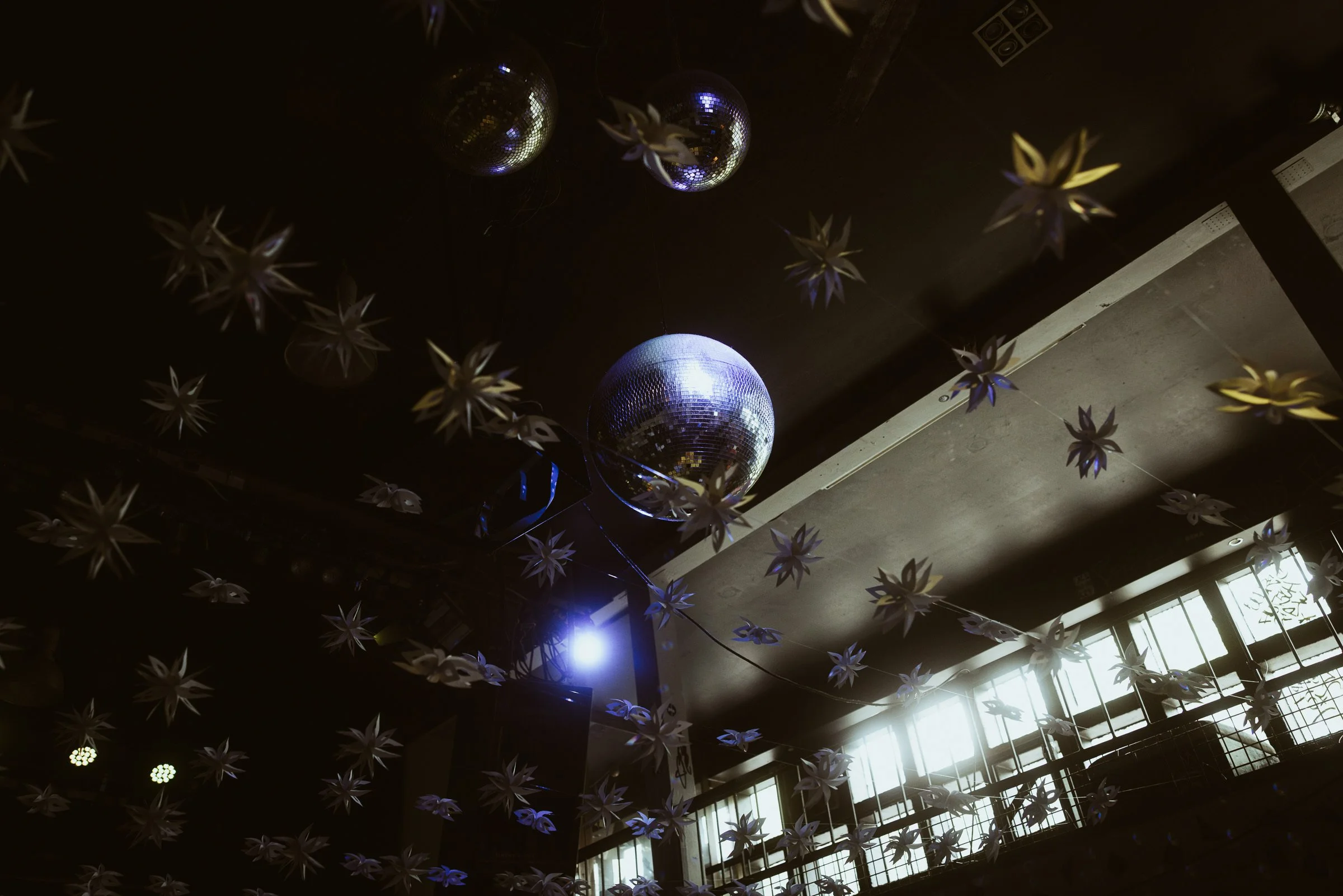 Dekorative Weihnachtssterne und Discokugeln hängen an der Decke eines dunklen Raumes, mit natürlichem Licht, das durch große Fenster auf der rechten Seite strömt.
