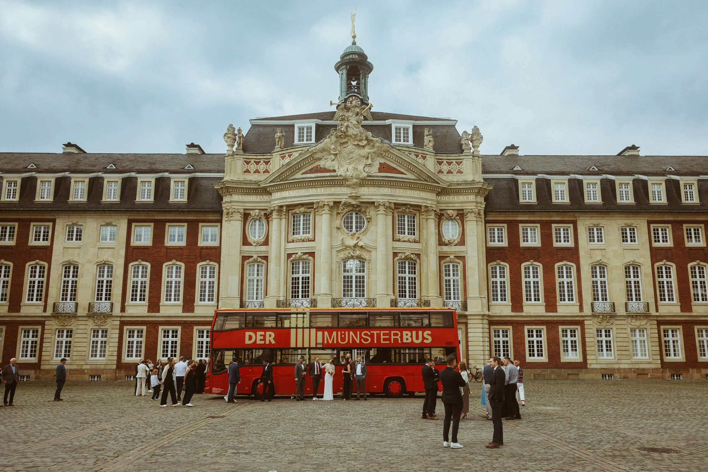 Historisches Gebäude mit barocker Architektur, darunter ein roter Doppeldeckerbus mit der Aufschrift "DER MUNSTERBUS" und Menschen, die vor dem Gebäude stehen, einige in stilvoller Kleidung.
