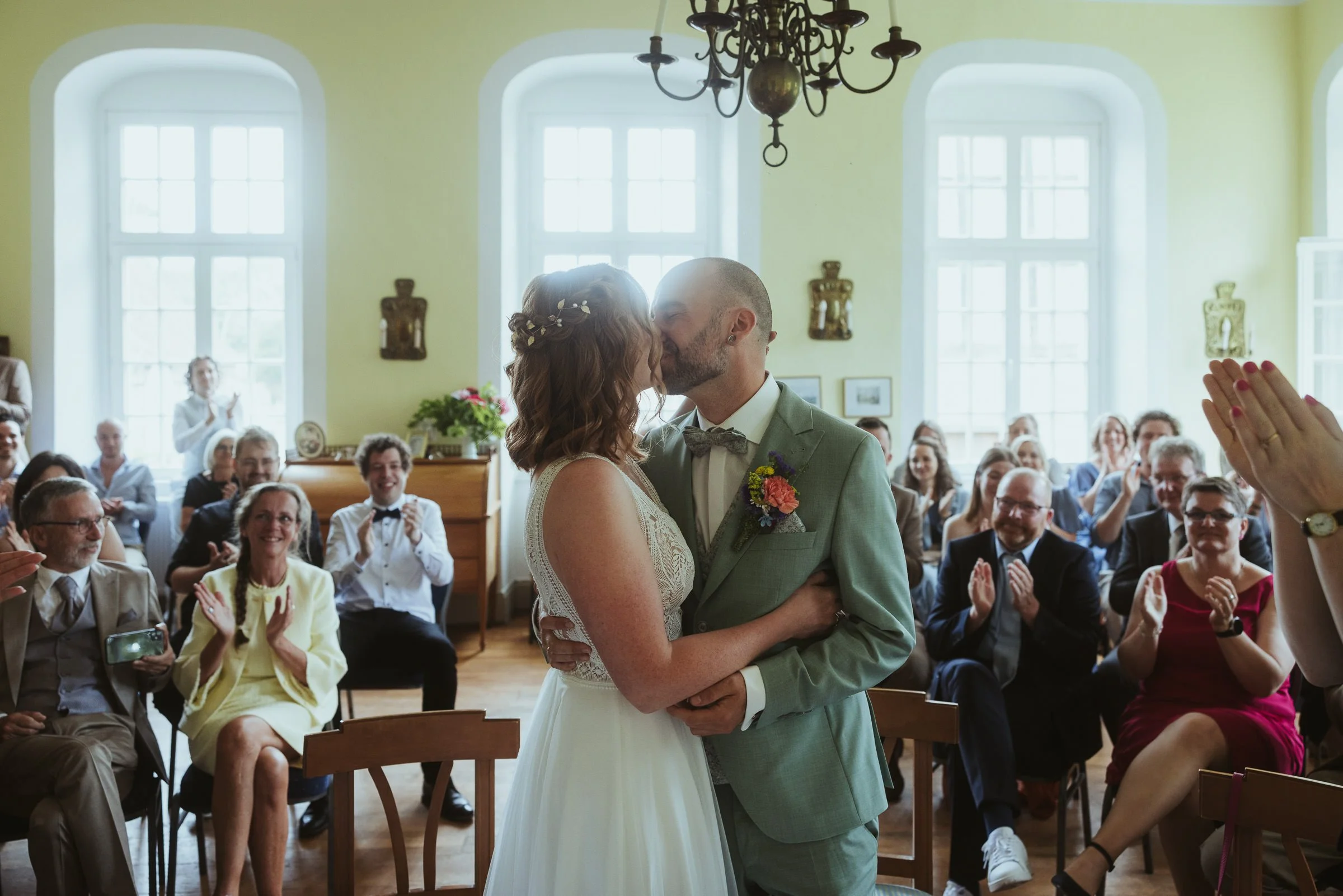 Ein Brautpaar bei ihrer Hochzeit im Saal, umgeben von jubelnden Gästen, die sich freuen und applaudieren.