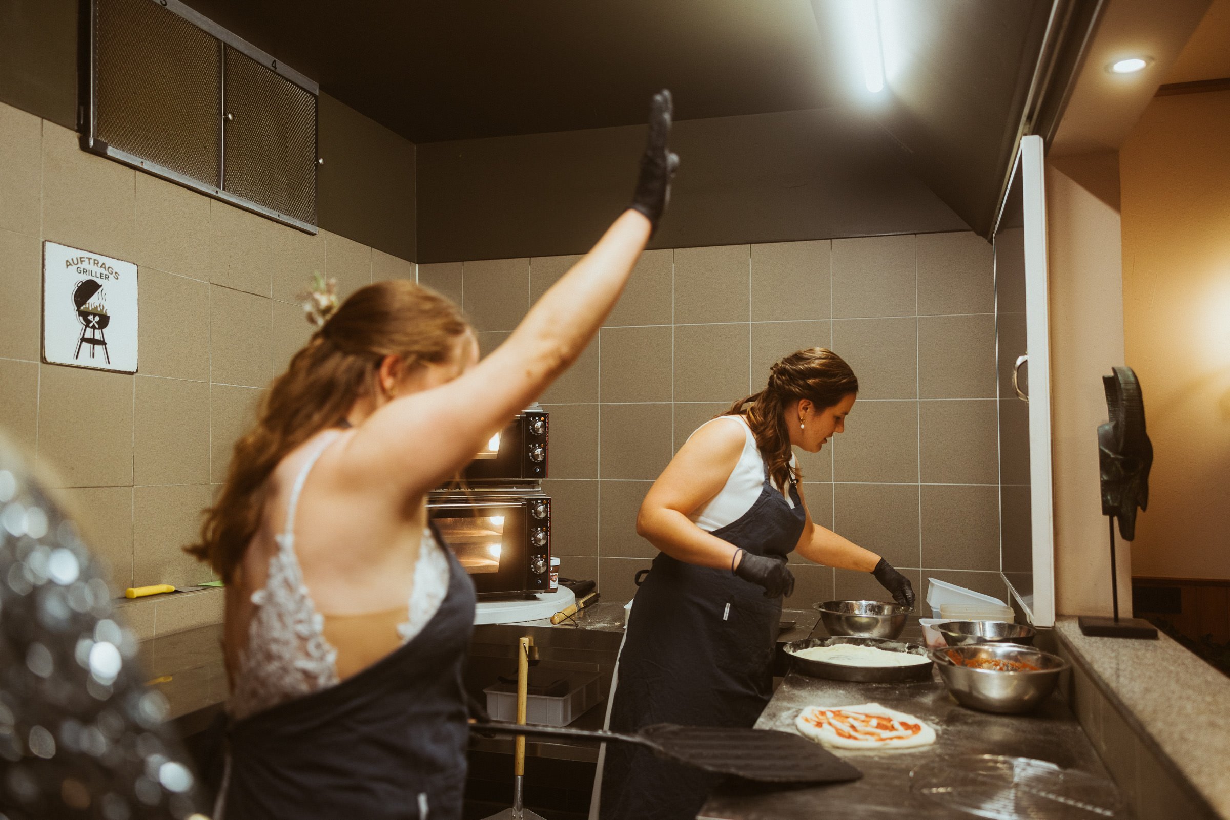 Zwei Frauen bereiten Essen in einer Küche vor, eine wirft eine Hand in die Luft, die andere schneidet Zutaten.