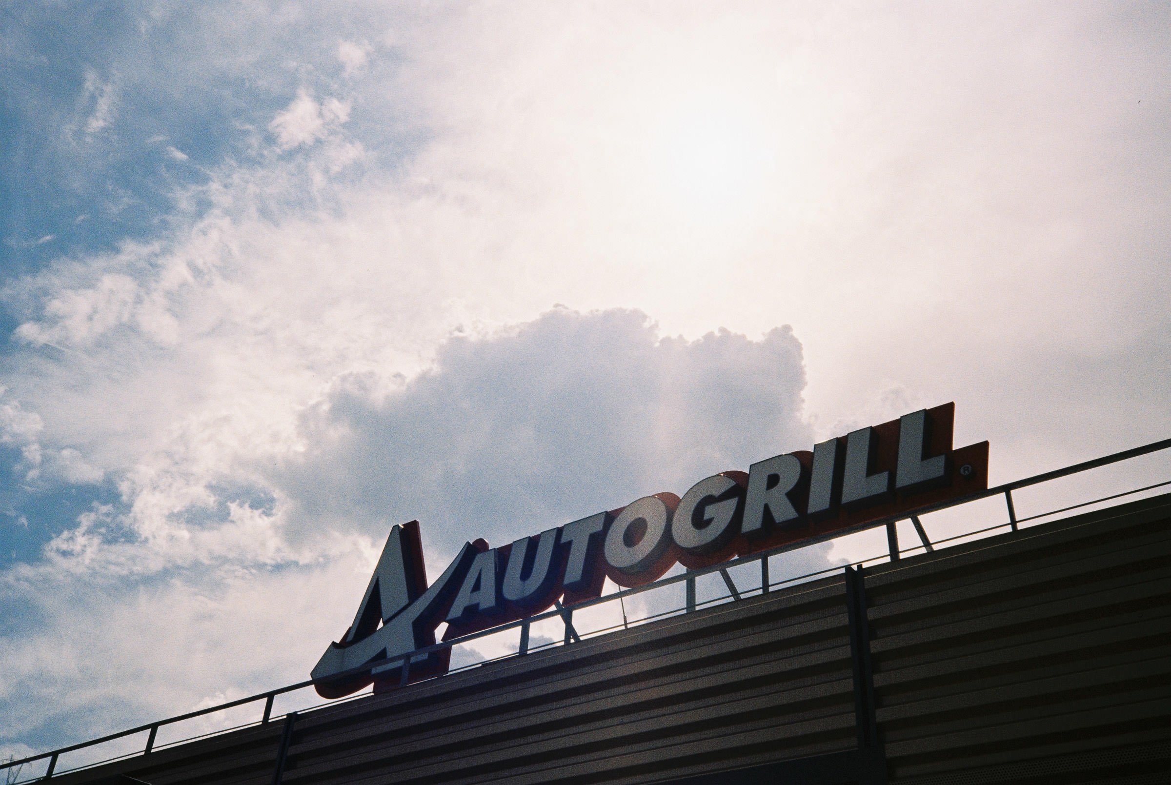Der Himmel ist teilweise bewölkt mit einigen kleinen Wolken, und im unteren Bereich ist das Dach eines Gebäudes mit einem großen, alten Unternehmensschild, auf dem die Wörter 'Auto Grill' zu erkennen sind.