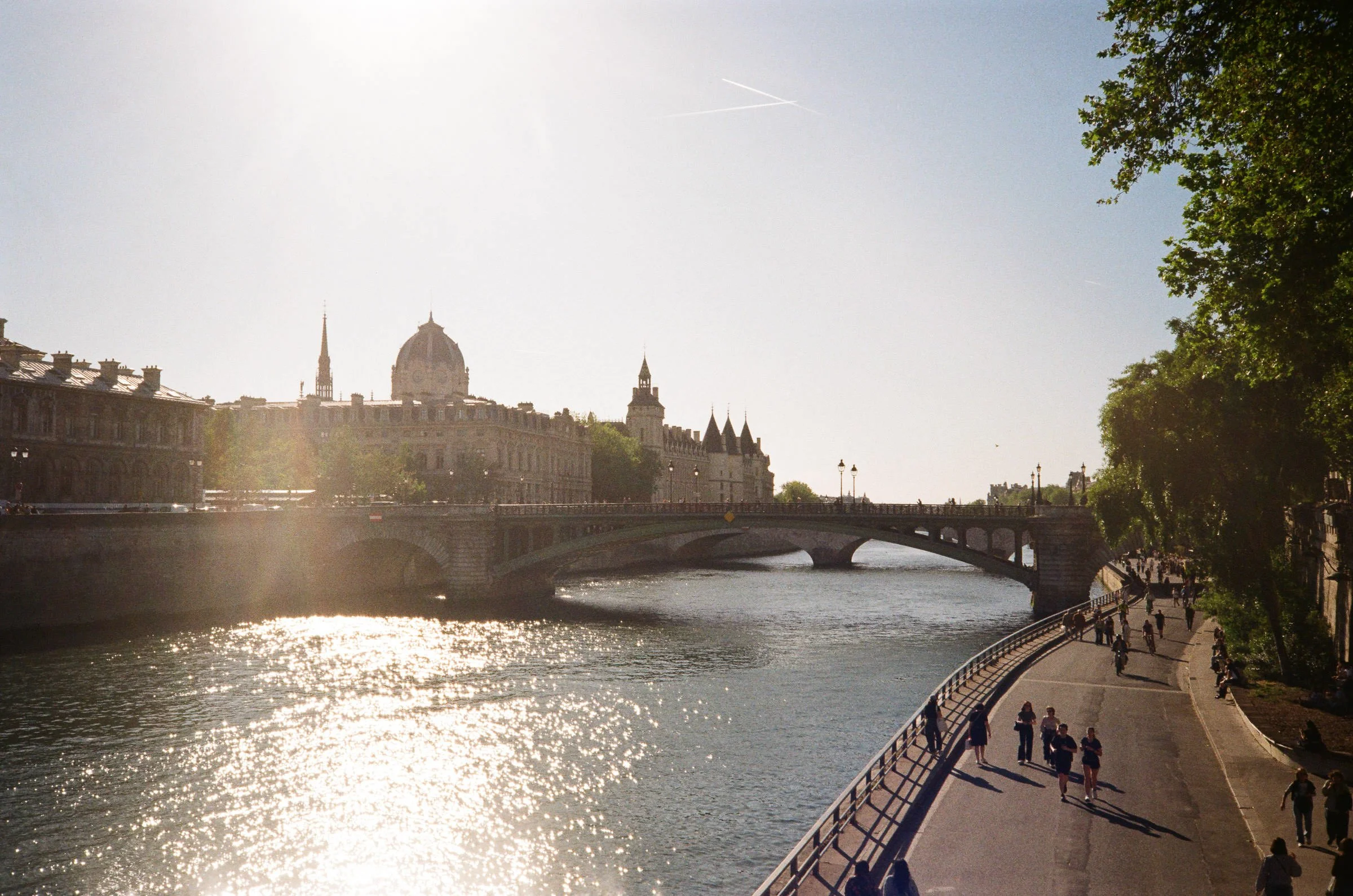 Blick auf die Seine in Paris bei Sonnenuntergang, mit Brücke und Passanten am Flussufer, historische Gebäude im Hintergrund und Bäume an der Uferpromenade.