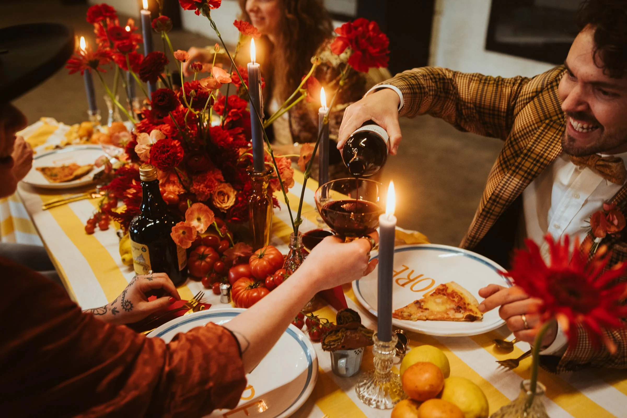 Menschen feiern an einem gedeckten Tisch mit roten Blumen, Kerzen und verschiedenen Speisen bei einem Abendessen.