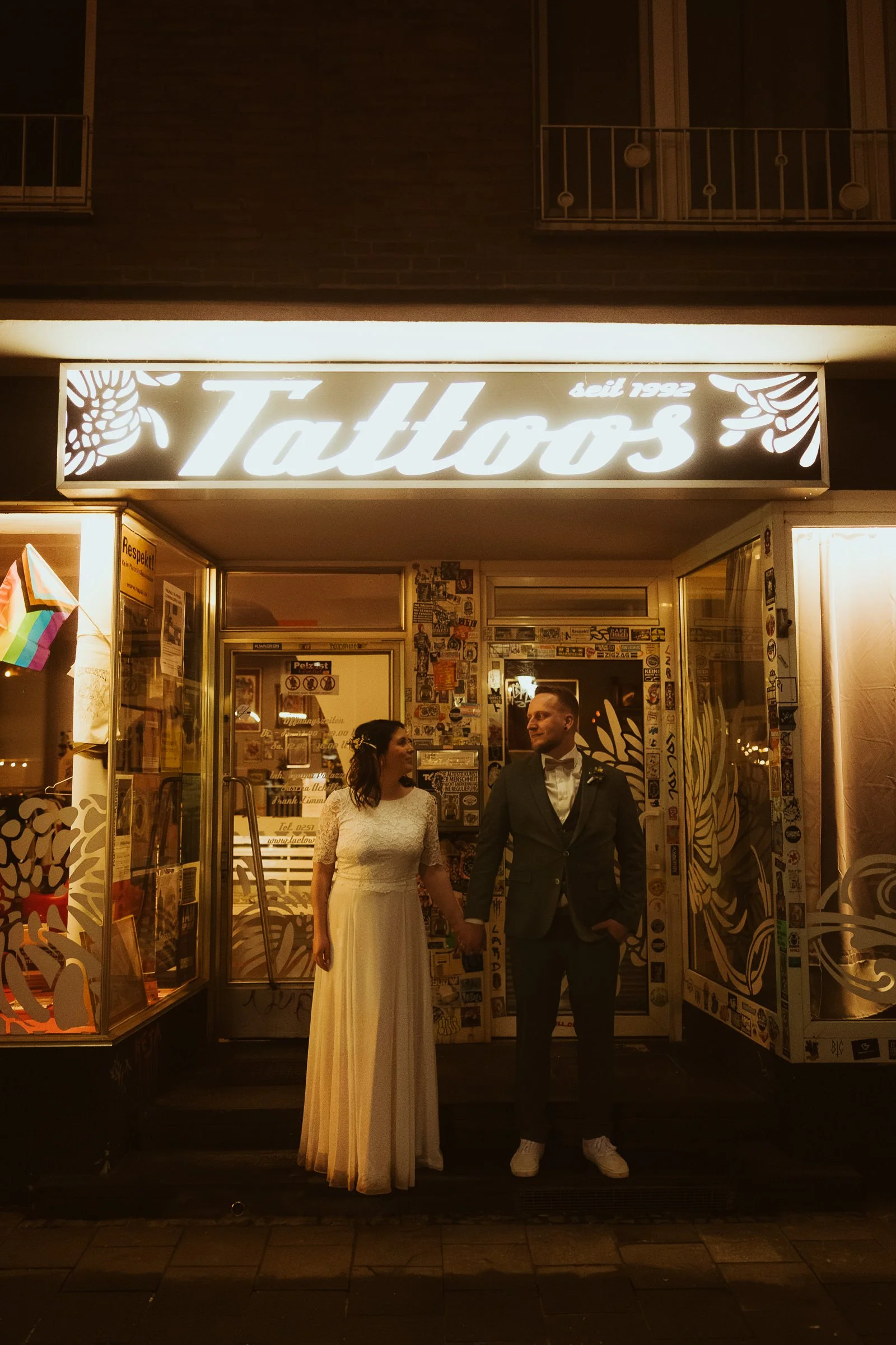 Hochzeitspaar hält Händchen vor einem beleuchteten Tattoo Studio am Hansaring in Münster.